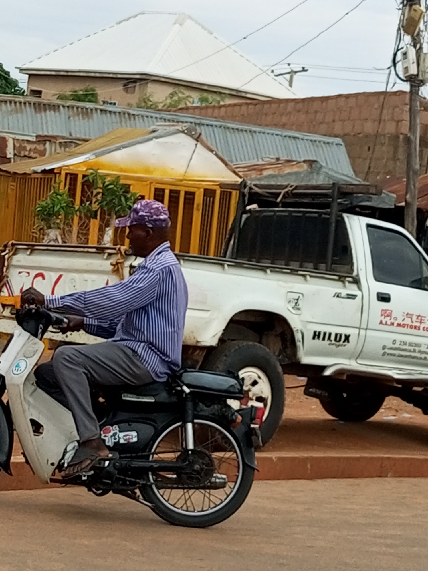 An eye-level, medium shot captures a Black man riding a white scooter on a street in an urban setting. He is wearing a purple patterned cap, a blue and white striped long-sleeved shirt, and grey pants. The man is looking to the left as he rides. Behind him, a white Toyota Hilux pickup truck is parked on the street. The truck has a black cage on the back and is displaying a logo with Chinese characters and "ALM MOTORS CO". The environment appears to be a developing urban area, with various buildings in the background constructed with corrugated metal roofs, brick, and light-colored siding. The sky is overcast.
