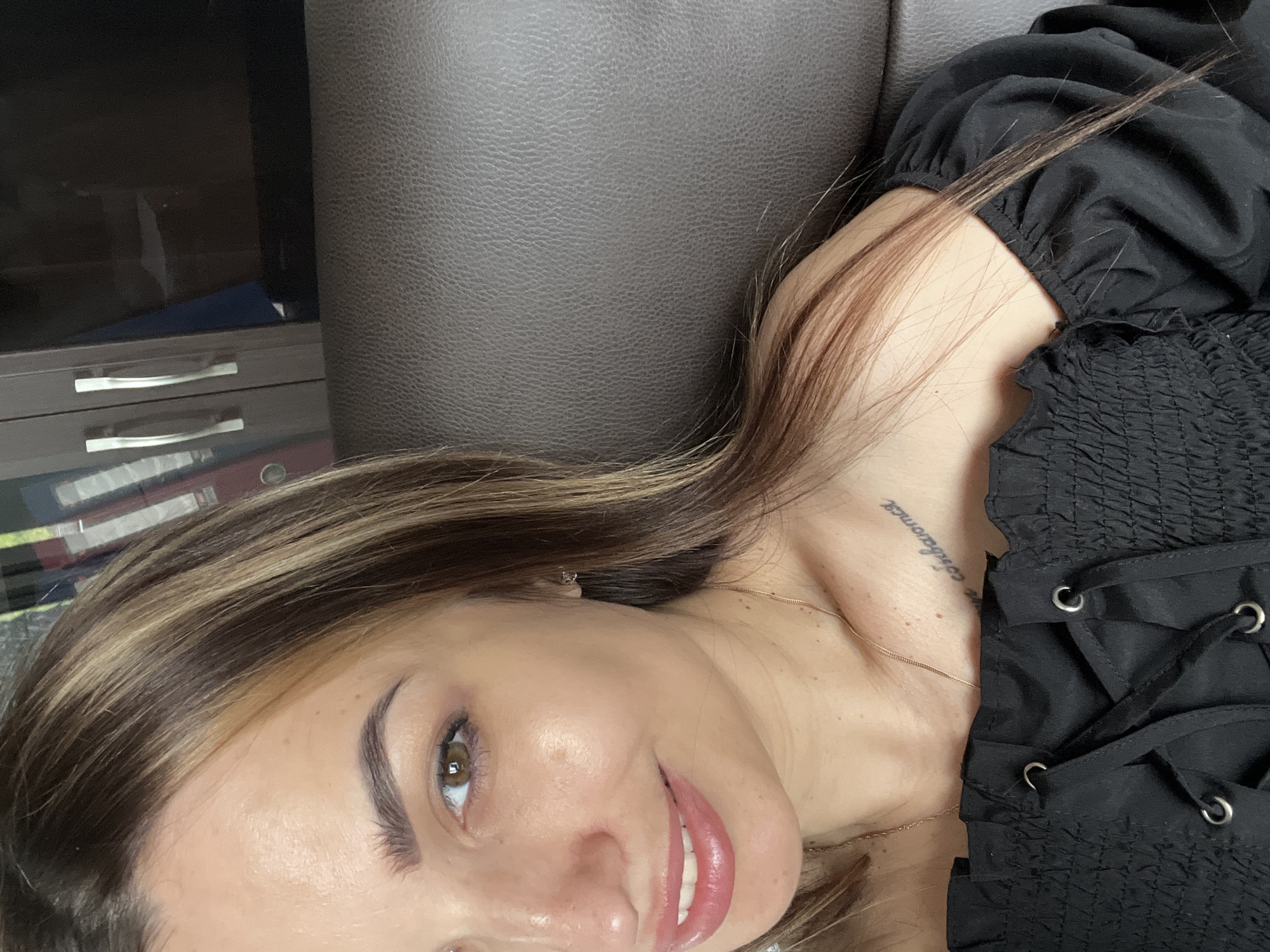 A close-up shot features a woman with light brown hair and blonde highlights, lying down with her head tilted to the right. She has light skin, brown eyes, and is smiling slightly, revealing her teeth. She is wearing a black top with a corseted design visible on the right side of the image. A gold necklace is visible around her neck, and a tattoo is present on her collarbone. The background consists of a dark brown leather-like surface, likely a couch or chair. A cabinet is visible in the background. The location is Unknown, Unknown.