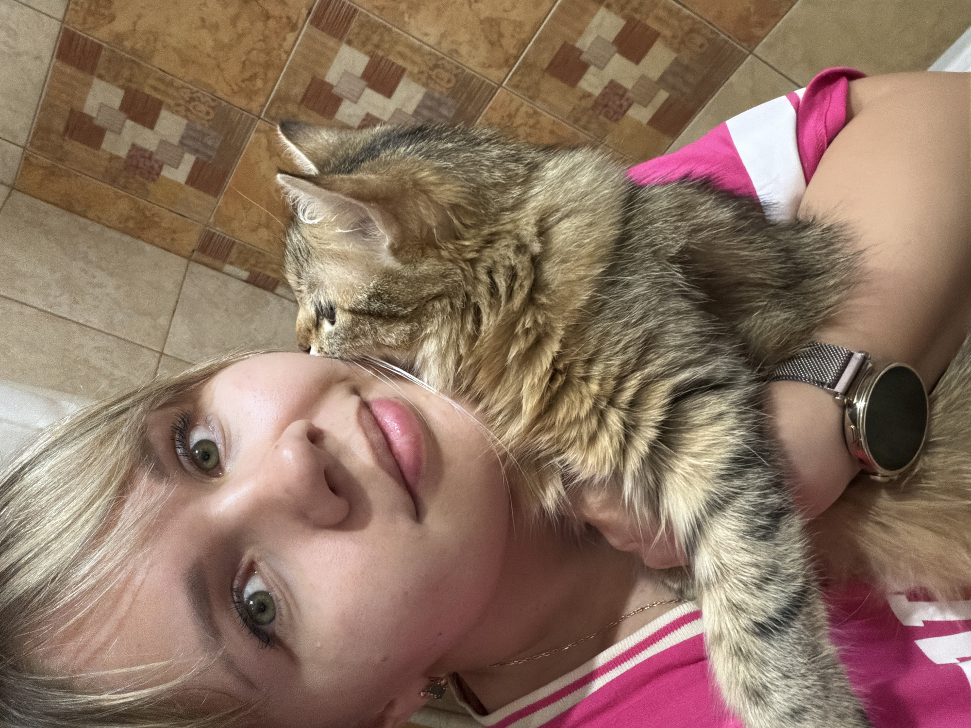 A young woman with light blonde hair, fair skin, and green eyes is holding a brown tabby cat close to her face in a selfie. She is wearing a pink and white striped shirt and a watch on her left wrist. The cat's head is near her forehead, and its tail is draped down her arm. The background includes a tiled wall in shades of brown and beige, and a white surface, possibly a pillow or bedding, is partially visible. The location appears to be indoors in an unidentified city.