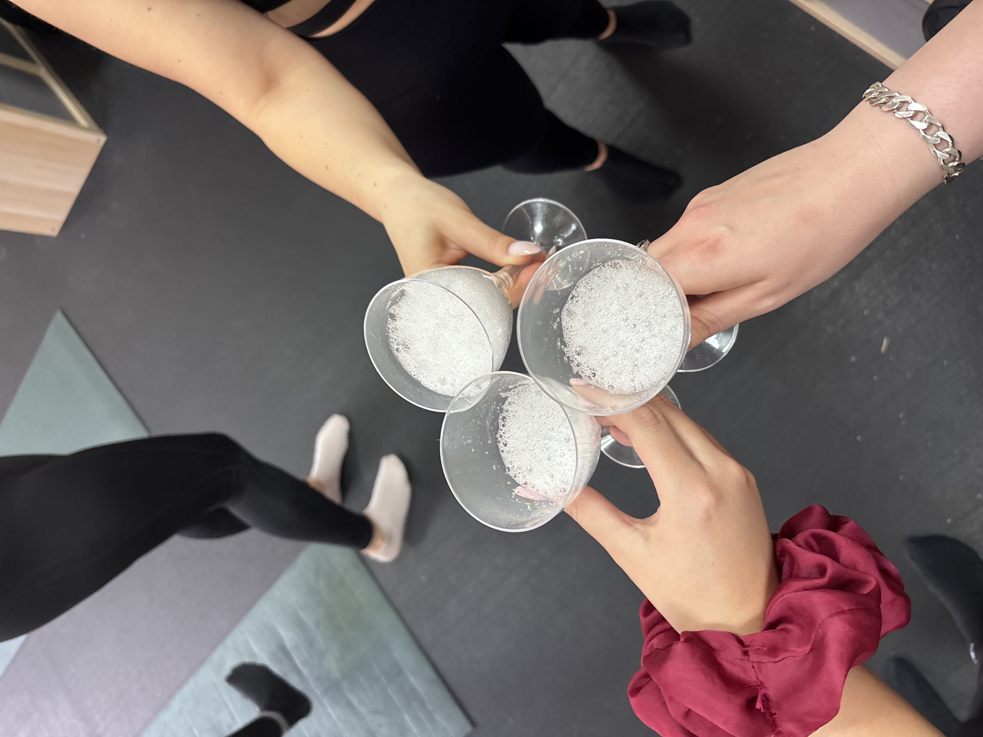 Four people in black leggings and socks/stockings stand in a circle, toasting each other with glasses of sparkling white wine. One person has a silver bracelet on their wrist and another has a red scrunchie. The location appears to be indoors on a black rubber mat. The city of Varna, Bulgaria, is not evident from the image.