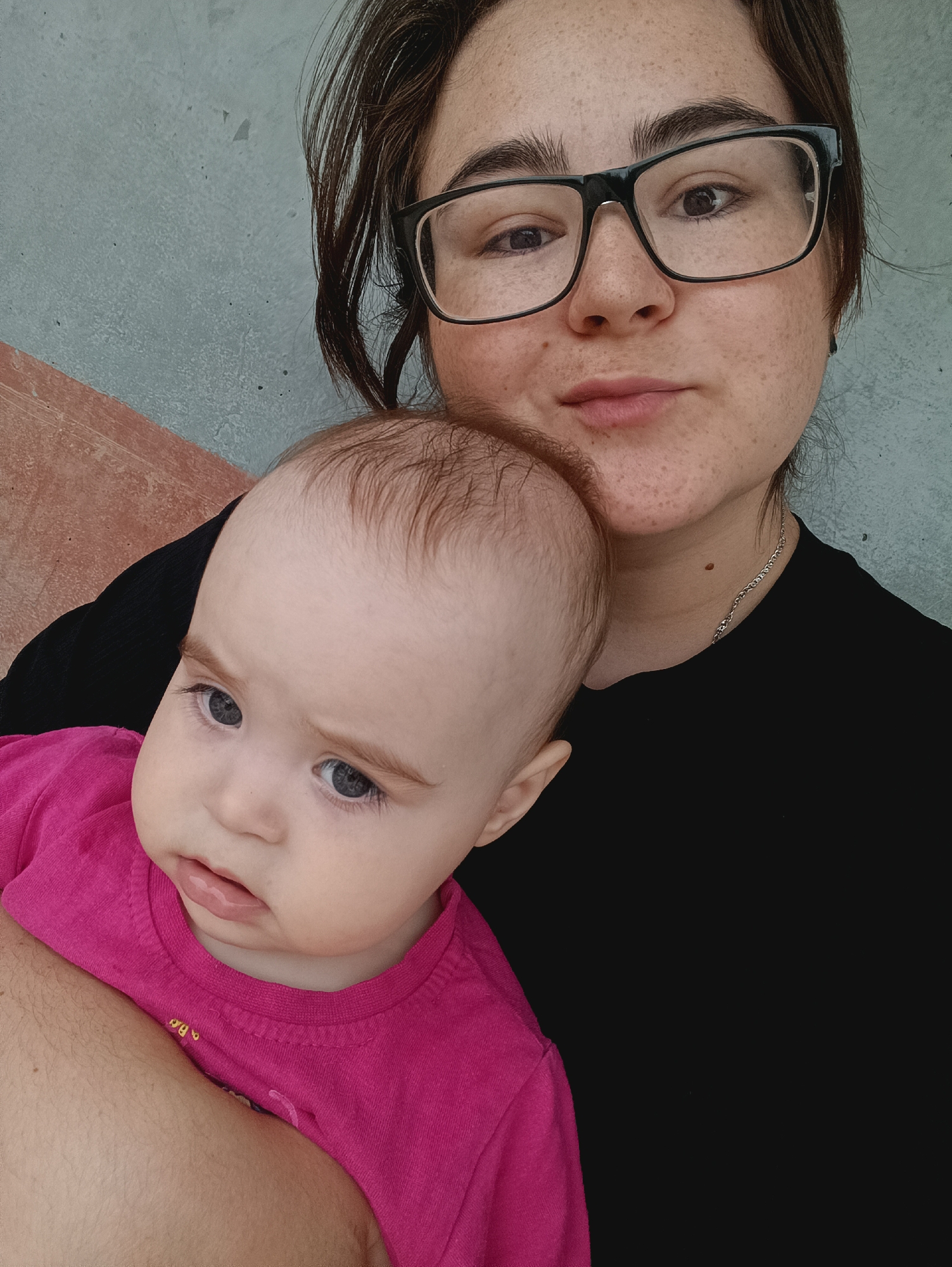 A medium shot selfie shows a young woman with dark hair and glasses holding a baby. The woman is wearing a black shirt, and the baby is wearing a pink shirt. They are against a textured wall with a diagonal line of lighter gray and a pinkish-orange area. The woman is looking towards the camera with a neutral expression, while the baby is looking slightly to the side with a curious expression. The background appears to be an outdoor urban setting, possibly a wall in the city of Sergiev Posad, Russia.