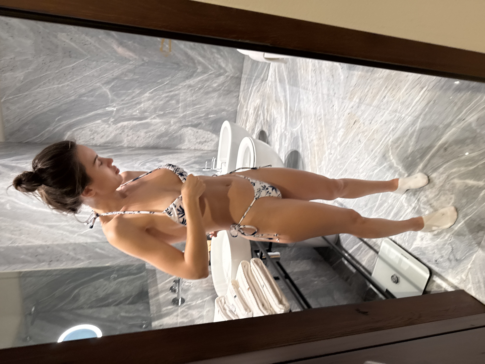 This full-shot image captures a female individual standing in profile within a modern bathroom with light grey marble walls and floor, accented by darker veining. The individual, with dark hair styled in a bun, is observed adjusting the top of a two-piece string bikini, which features a white background with blue and black patterns. She wears white, sock-like foot coverings.
The bathroom is furnished with white contemporary fixtures. On the left, a white circular basin with chrome faucets is visible, below which a stack of neatly folded white towels rests on a shelf. A glass shower screen behind the basin reflects a circular illuminated light source. To the right of the basin, a white wall-mounted toilet is present, followed by a matching wall-mounted bidet, both equipped with chrome fixtures. A white digital bathroom scale is situated on the marble floor in the lower left quadrant. Dark wooden frames are visible along the left and right edges of the scene.