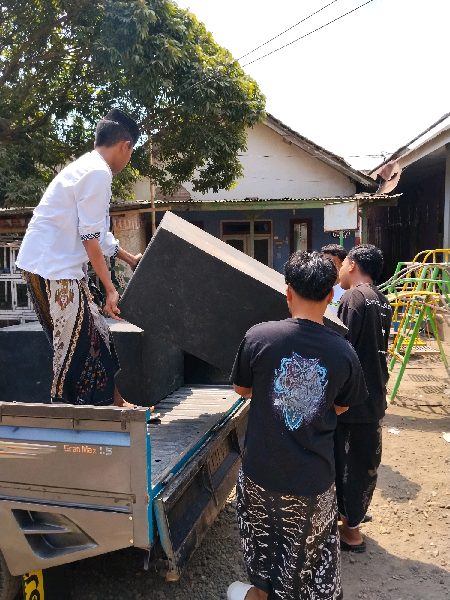 In Panggungan, Indonesia, under a bright, clear sky, a group of four young men are actively engaged in loading large, black, rectangular objects, likely speakers, into the bed of a light blue pickup truck labeled "Gran Max 1.5". The truck bed is lined with what appears to be a rough, grey material. One man, wearing a white long-sleeved shirt and a patterned sarong, is positioned on the edge of the truck bed, appearing to guide or push one of the objects. Two other men, dressed in black t-shirts and patterned sarongs, stand behind him, also appearing to be involved in the loading process. The man in the foreground, facing away from the camera, wears a black t-shirt with a detailed owl graphic on the back. The fourth man, also in a black t-shirt with writing on the back and a sarong, is partially obscured. The environment suggests a casual outdoor setting, possibly a residential area, with a building with a tin roof visible in the background, a large tree with dense green foliage to the left, and a playground structure with yellow and green frames to the right. The ground is unpaved and appears to be composed of dirt and gravel. The overall scene depicts a collaborative effort to move heavy equipment.