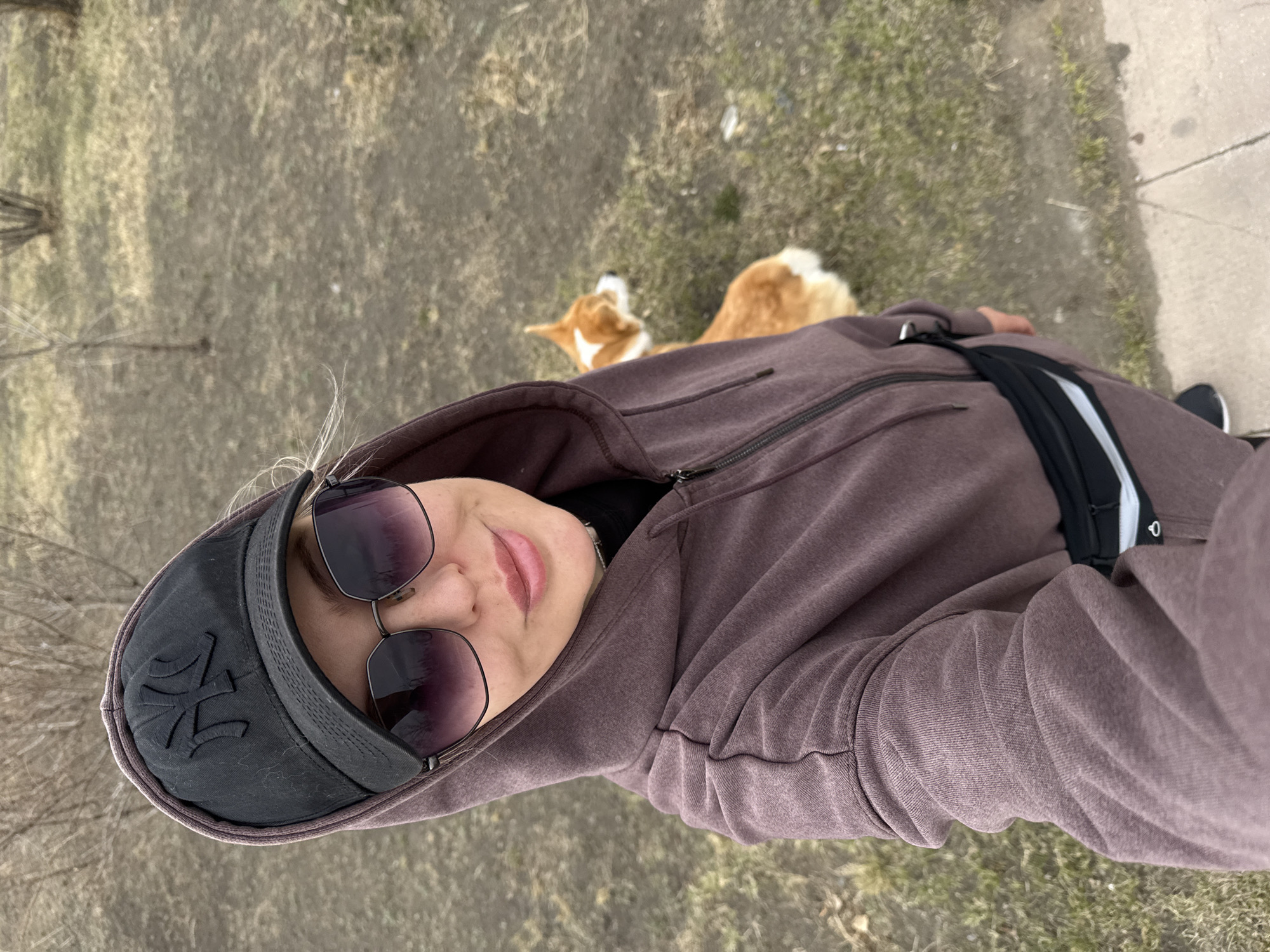 A medium shot shows a person and a dog outdoors. The person, wearing sunglasses, a dark baseball cap with an embroidered logo, and a hooded brown jacket, appears to be holding the camera for a selfie. The dog, a light brown and white breed, is lying on the ground in the background. The ground is covered in dry grass and patches of bare earth. A paved surface is visible on the right edge of the frame. This is speculated to be the location in the city of Borzya, Russia.