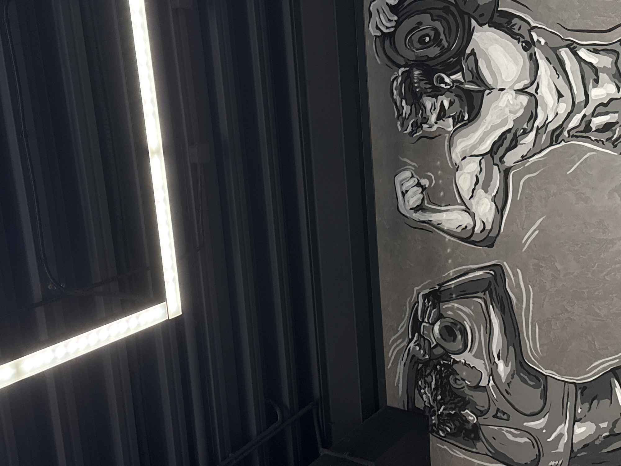 The image shows a portion of a building's interior with a focus on a mural and ceiling lighting. The mural depicts a muscular figure lifting weights and a separate figure below it, both rendered in grayscale with bold outlines. The ceiling is constructed of dark, corrugated metal panels. An LED light fixture in the shape of an incomplete square is attached to the ceiling and is prominently lit. The location in the city of Votkinsk, Russia, is unknown; no features in the image indicate the area.
