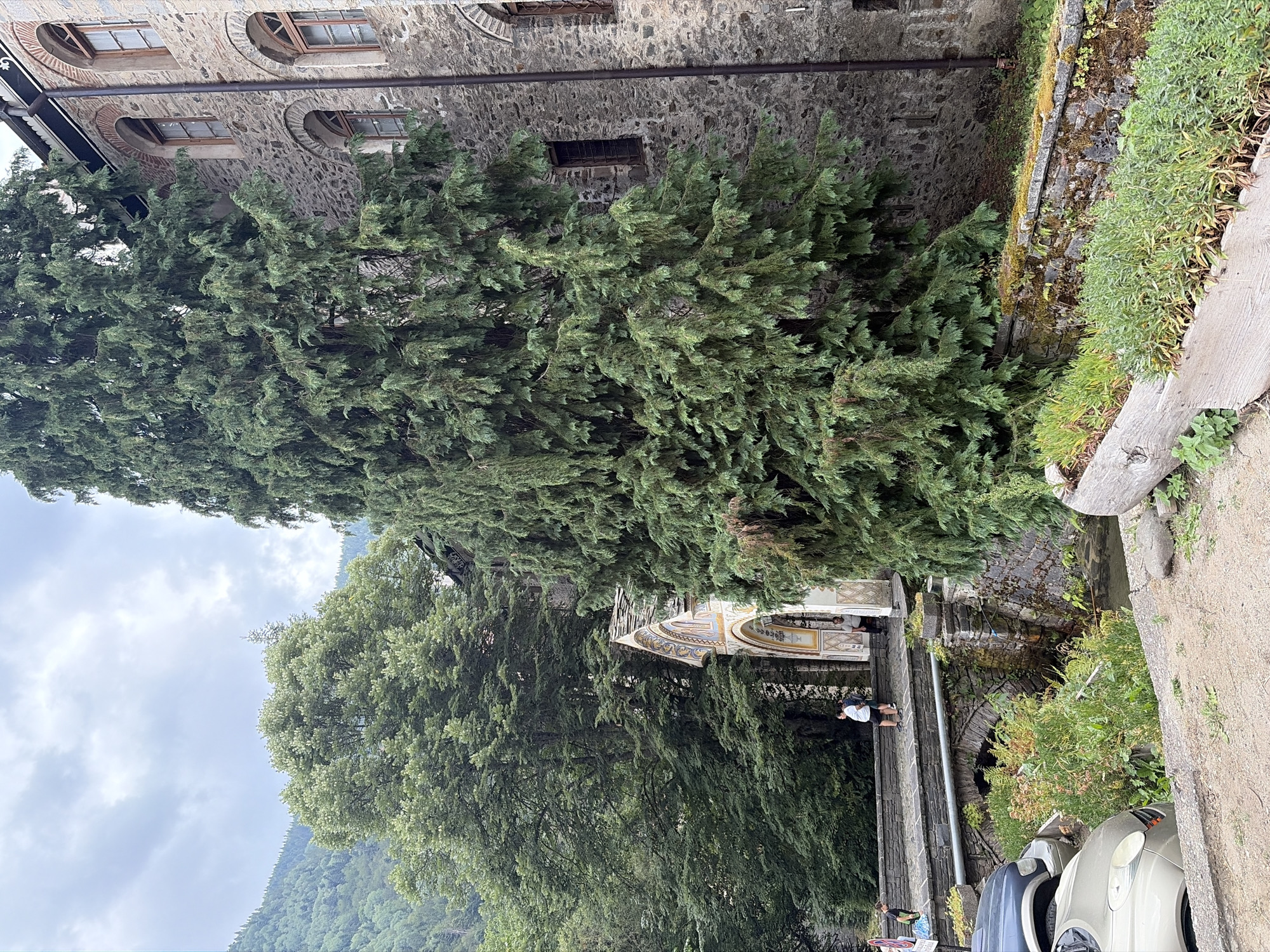 The image depicts a stone building with arched windows, overgrown with a large evergreen tree in the foreground. A bridge crosses a stream, leading to an arched entryway decorated with intricate mosaics. Several people are visible on the bridge and near the entrance. The surrounding environment includes lush green trees and vegetation, with a mountainous landscape in the background. The scene appears to be the exterior of a historic religious site, likely the Rila Monastery in Bulgaria, given the architectural style and natural surroundings. The sky is overcast.
