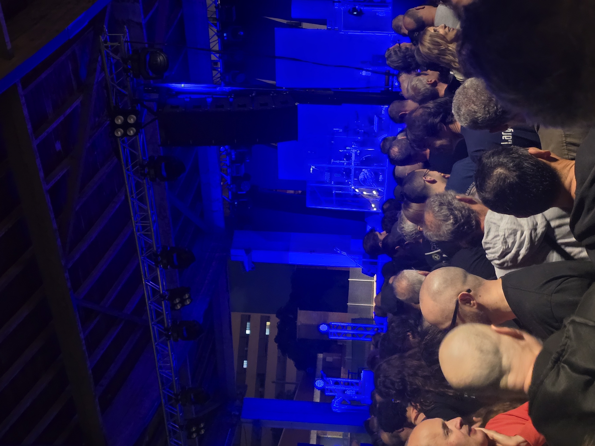 This image captures a nighttime event, likely a concert or performance, in Cornellà De Llobregat, Spain. The scene is illuminated predominantly by intense blue stage lighting, with a dark wooden ceiling structure and suspended metal trussing visible at the top. Numerous stage lighting fixtures and large line array speaker systems are mounted on the trussing.
In the background, a raised stage area is set with various equipment, including a partially visible drum kit on the left, vertical light structures, and large, rectangular blue-lit backdrops. Beyond the stage, an urban environment at night is discernable, featuring a multi-story building with illuminated windows and a dark, possibly vegetated, elevated area.
The foreground and midground are occupied by a dense crowd of approximately 50-70 individuals, primarily adults, standing and oriented towards the stage. The majority are seen from behind or profile, consisting largely of men, many of whom are bald or balding, and a smaller number of women. Clothing is predominantly casual and dark, with one man in the far left foreground wearing a red shirt. A woman with long blonde hair in the right midground is smiling. One individual's black shirt displays partially legible text, possibly "Lary." The crowd appears to be observing the stage, with minimal active interaction visible.