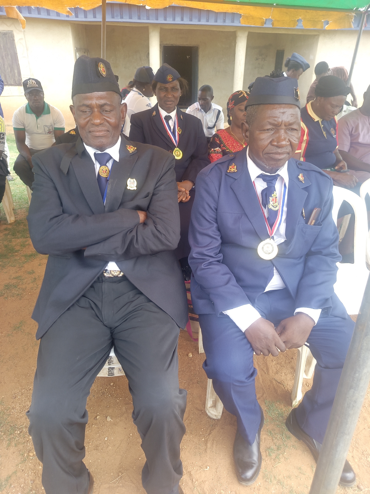 In an outdoor setting in Akwanga, Nigeria, two older men sit side-by-side on white plastic chairs. Both wear dark-colored, formal military-style uniforms with peaked caps adorned with insignia. The man on the left has a black uniform and is sitting with his arms crossed. The man on the right is wearing a blue uniform and has a large silver medal around his neck. Other individuals are seated behind them in the background, also on plastic chairs. A building with a light-colored exterior is visible in the background. The ground is reddish-brown soil. A canopy provides partial shade overhead.