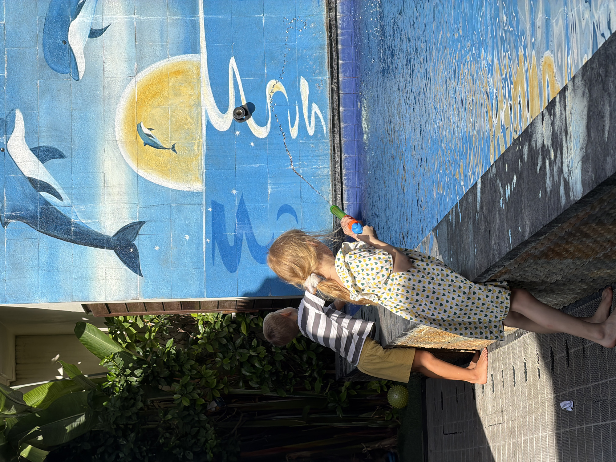 Two children are near the edge of a pool. One child, with blonde hair, holds a water gun and appears to be aiming it into the pool. The other child stands nearby. The pool's wall features a mural with dolphins and a sun on blue tiles. The pool is located in Kuta, Indonesia.