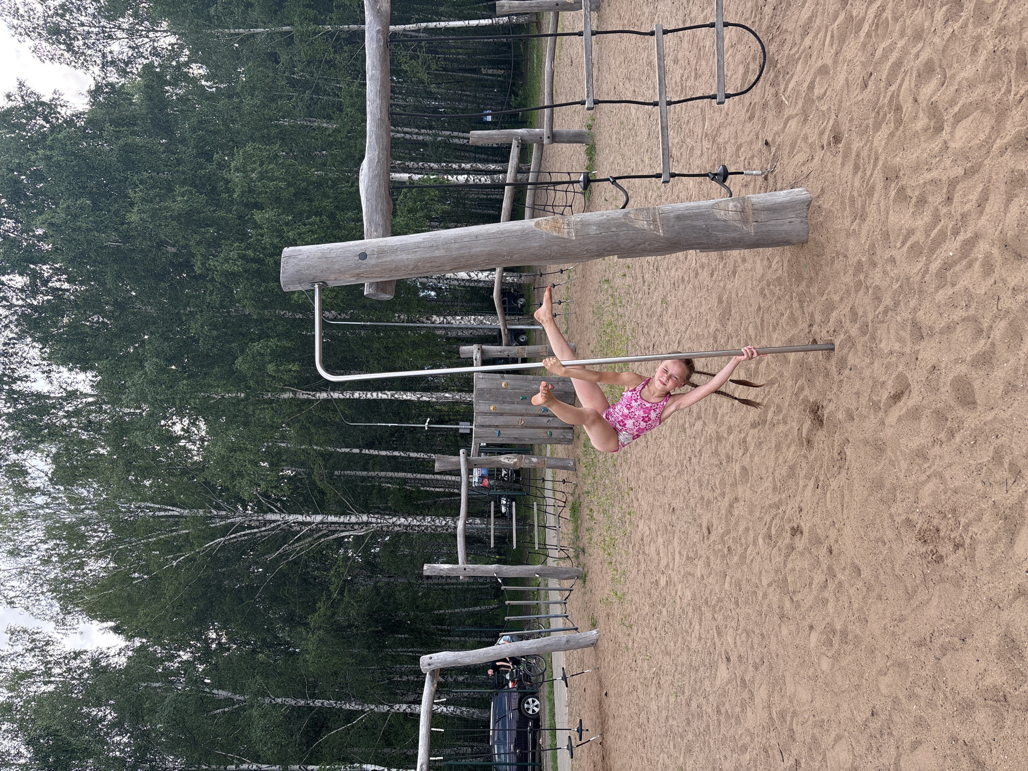 A young girl in a pink floral swimsuit is performing a gymnastic move on a metal bar suspended from a wooden playground structure. She is holding onto the bar with one hand and her legs are extended upwards. Her body is arched, with her head tilted back and a smile on her face, suggesting she is enjoying herself. The playground is made of natural wood and metal, set on a sandy ground, with a backdrop of lush green trees. In the background, a black SUV and a person in a wheelchair are visible near the edge of the playground, indicating a public park or recreational area. The lighting suggests it is daytime. The image is oriented vertically, with the girl appearing to be hanging from the top of the frame.