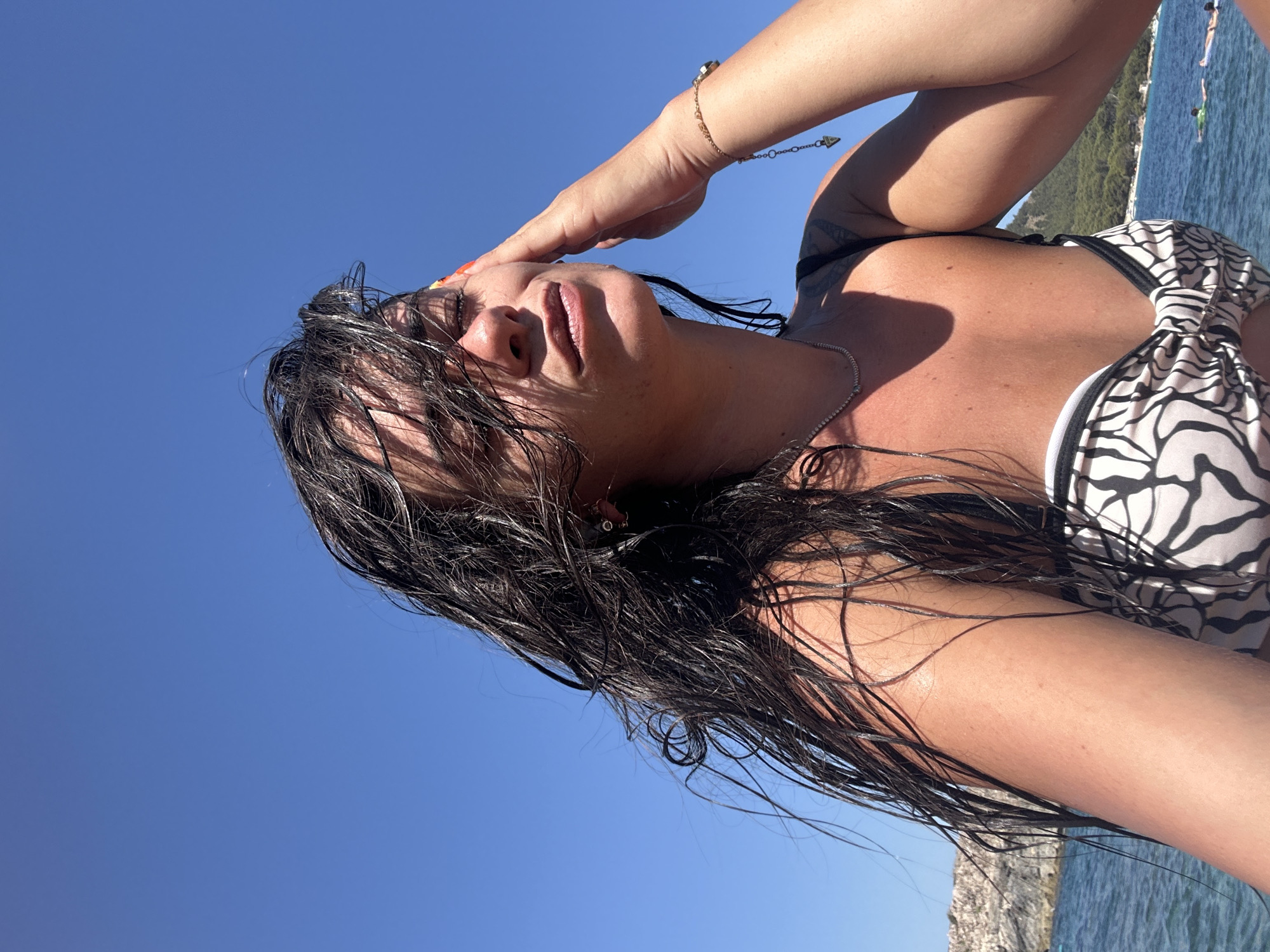 A woman with wet, dark hair is seen from the chest up, against a clear blue sky. She is wearing a black and white patterned bikini top and a delicate necklace. Her right hand is touching her forehead. The background shows a body of water with a rocky shore to the left and a green hillside on the right. The scene appears to be at Cala Lliteres, Spain.