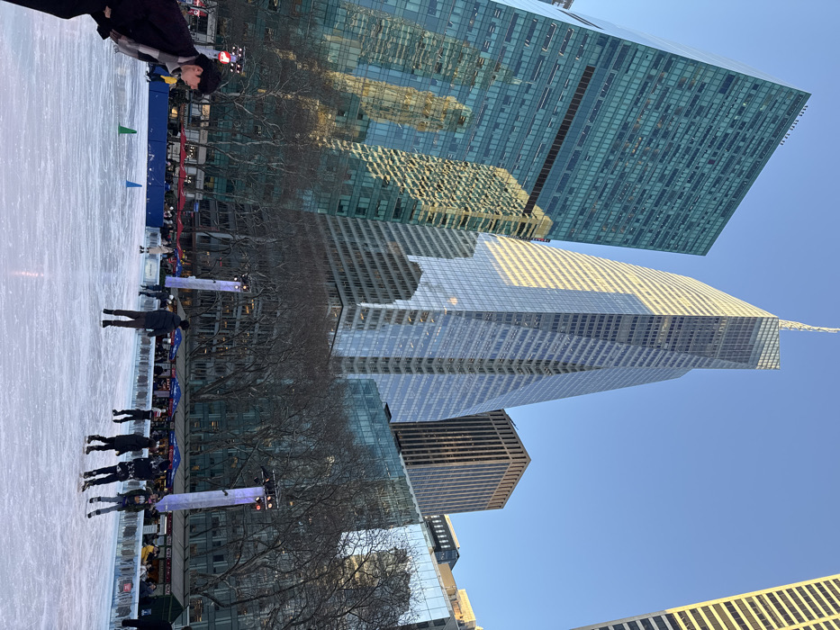 A winter scene showcases an ice rink bustling with skaters. Towering skyscrapers frame the view, reflecting light under a clear blue sky. Bare trees outline the rink's perimeter, with a small crowd observing the activity. The composition captures urban recreation against the backdrop of modern architecture.