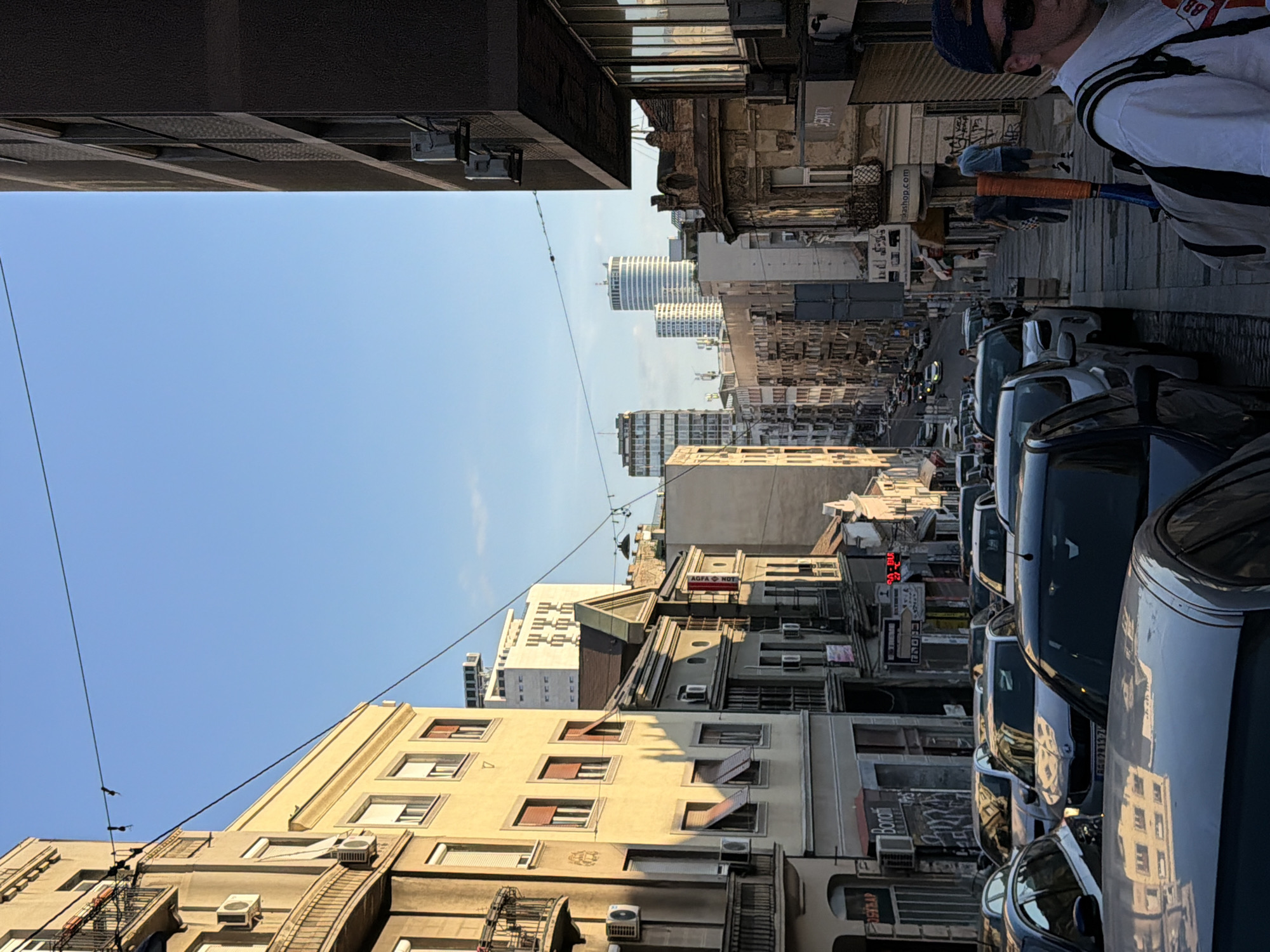 The image shows a street in Belgrade, Serbia, lined with parked cars. Buildings of various heights and architectural styles flank the street, with taller modern buildings visible in the distance. A man with sunglasses and a tennis racket walks on the sidewalk. Utility wires stretch across the sky. The sky is clear and blue.