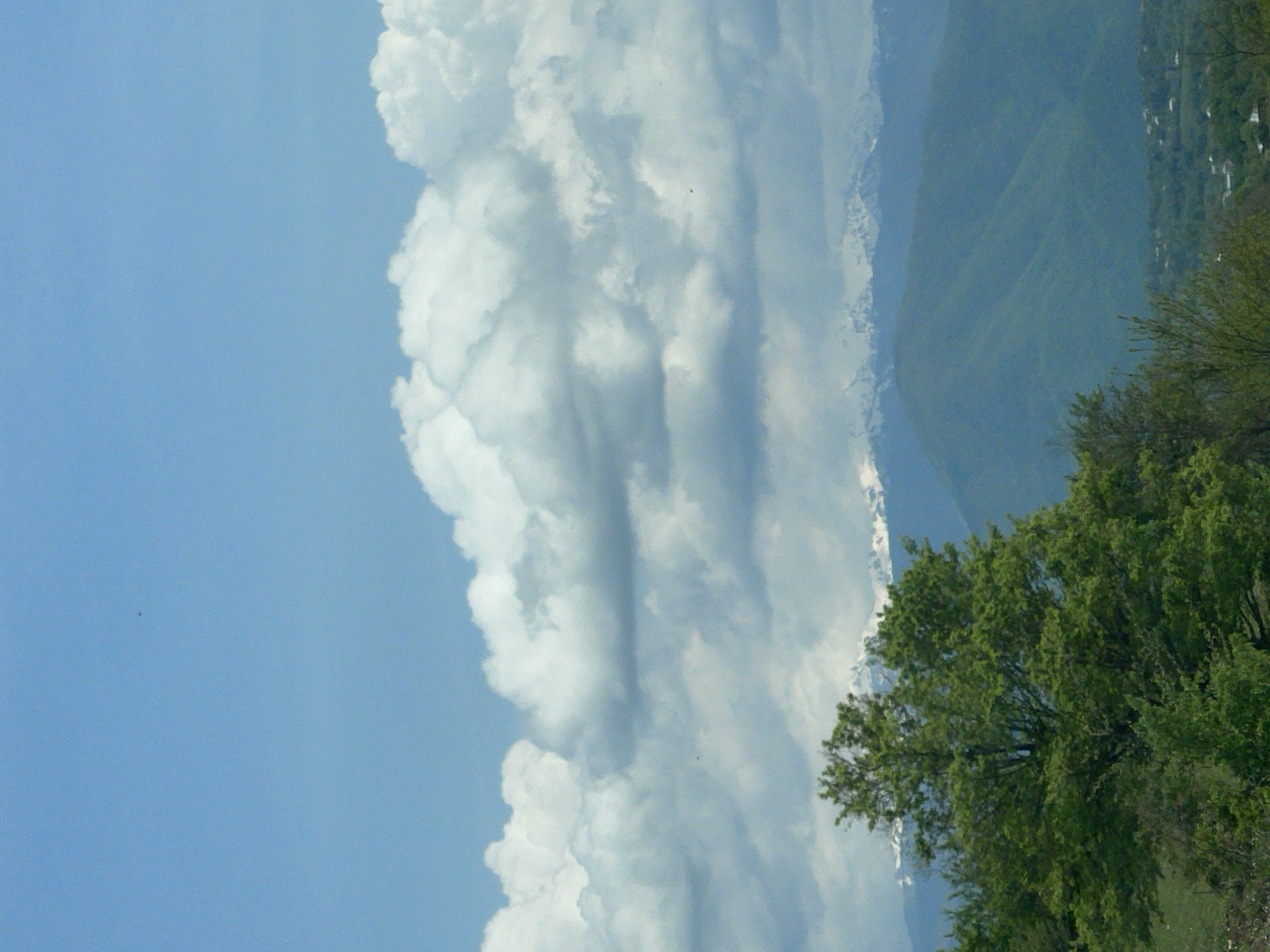 The image shows a sky with a large cumulus cloud formation taking up a significant portion of the scene. The sky is blue with varying shades, suggesting different atmospheric conditions. In the background, there's a mountain range, with snow-capped peaks partially visible. To the right of the frame, there are trees and vegetation. It cannot be determined that the location is Chiatura, Georgia.