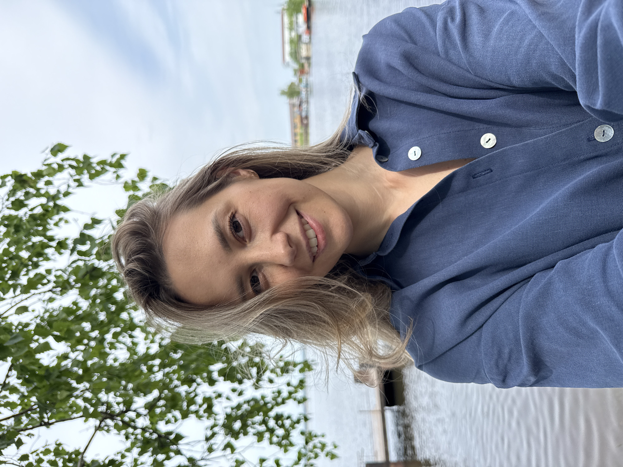 A fair-skinned woman with blonde hair is smiling at the camera while wearing a blue button-down shirt. The photo was taken outdoors near the water, possibly a lake or river. In the background is vegetation; on the water, there is a building. The image was taken in the city of Chaikovsky, Russia.