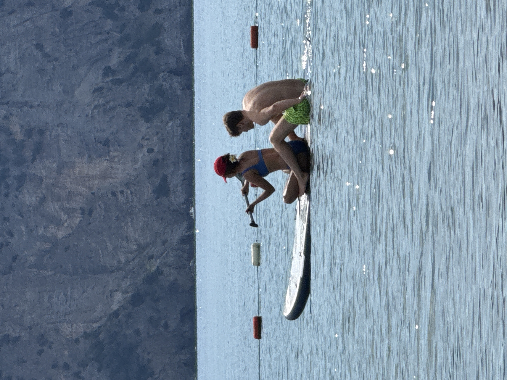 Two individuals are on a paddleboard in the water. One person, wearing a blue bikini and a red baseball cap, is sitting and holding a paddle. The other person, a male, is sitting behind them, facing away from the camera. He is wearing green patterned shorts. There are red and white buoys floating in the water behind them. In the background, a rocky mountain range is visible. The scene appears to be a coastal area, possibly near Prijevor, Montenegro, given the water and mountain landscape.