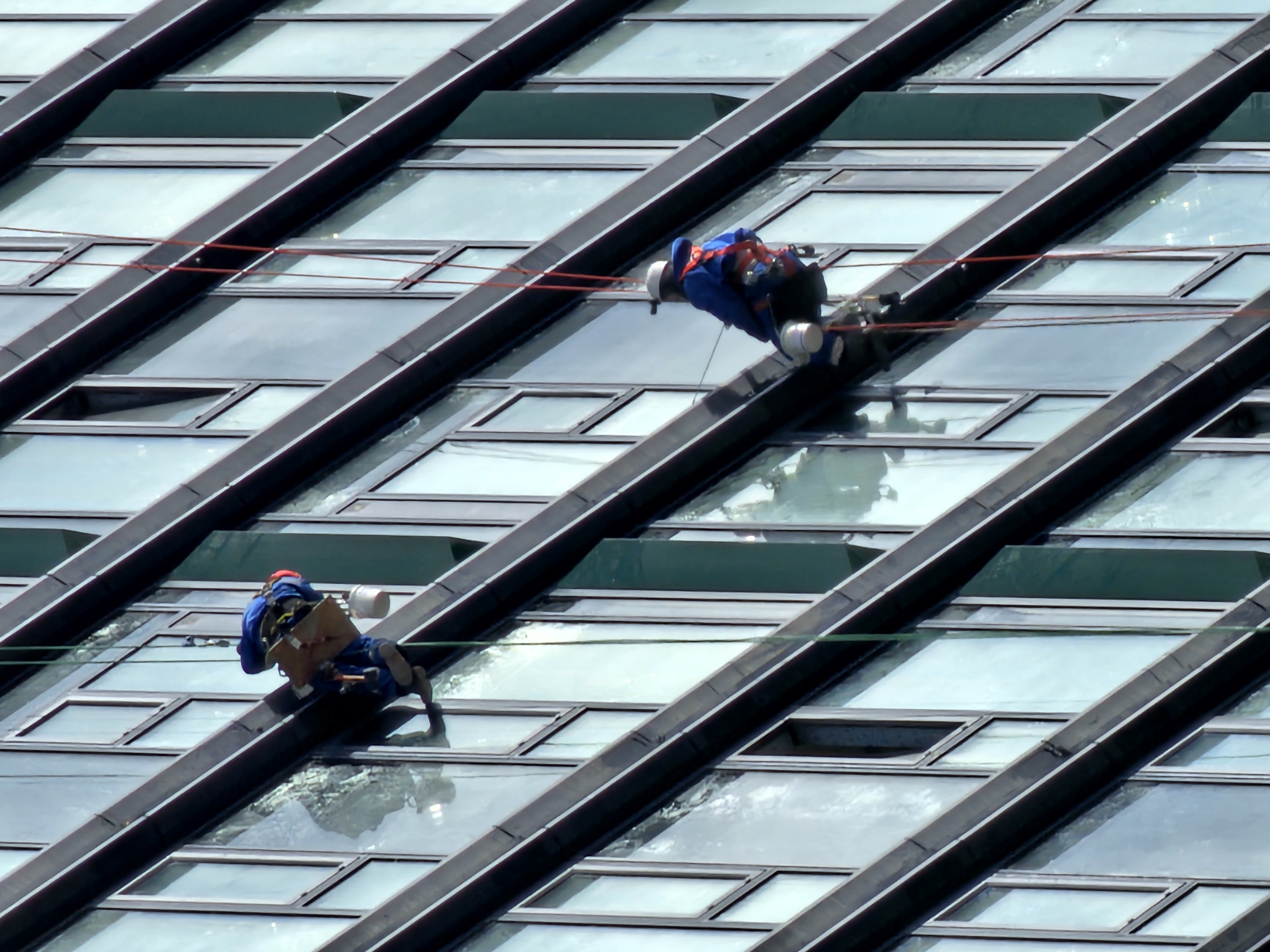 The image depicts two window washers suspended on the exterior of a modern glass-clad building. The building's architecture is characterized by angled or slanted window panels with dark frames, creating a geometric pattern. The washers are attached to ropes and harnesses, seemingly performing maintenance work. One is situated higher than the other. The upper washer wears a blue suit with a white helmet. The lower washer has a red helmet. Both are carrying equipment necessary for the job. The environment is bright, suggesting a sunny day. The building design suggests an urban setting, presumably in Moscow, Russia.