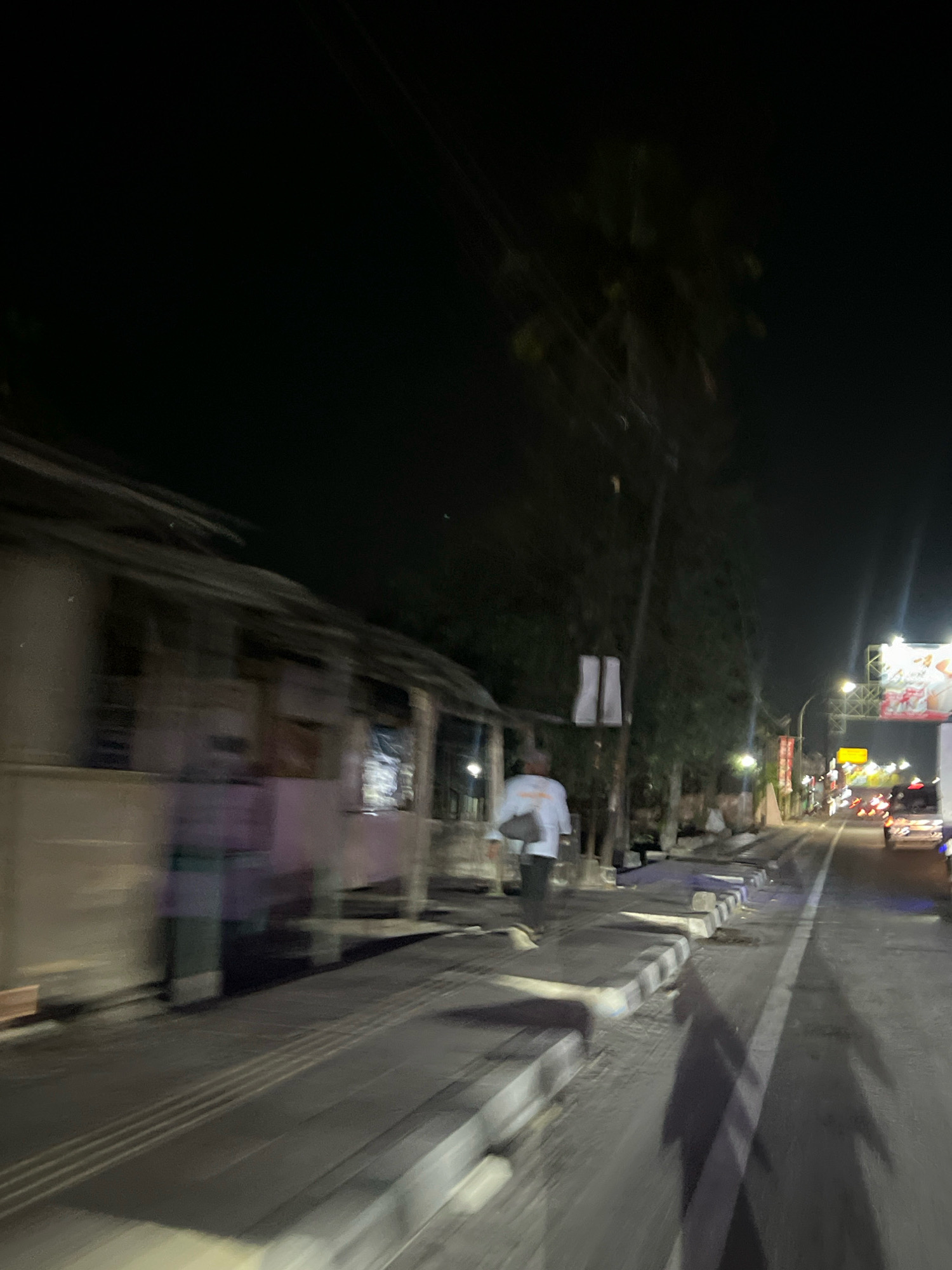 The image depicts a street scene at night in Sudimoro, Indonesia. A person in a white shirt and dark pants is walking away from the camera on a sidewalk. The sidewalk has a textured pattern and is bordered by a road with active vehicle traffic, indicated by visible headlights and taillights. On the left side of the image, a blurred building structure is present. Trees and streetlights line the street, contributing to the nighttime ambiance. The scene appears to be a commercial or residential area with visible signage and lighting.