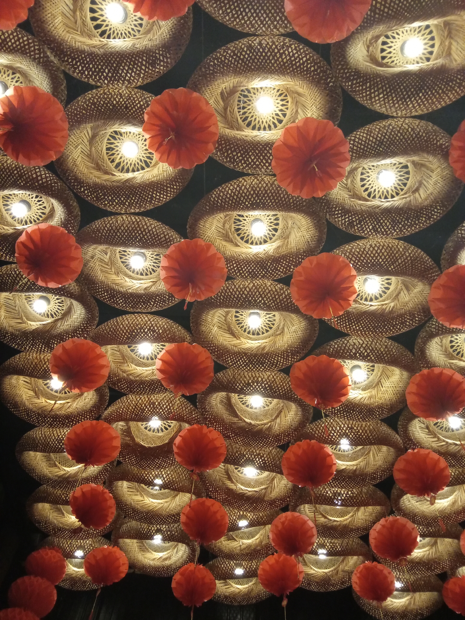 A low-angle shot shows a ceiling arrangement composed of woven, circular lampshades, interspersed with red paper lanterns. The lampshades have a light-brown color and a woven texture. The lampshade centers emit a bright light. Red paper lanterns are arranged in a grid pattern throughout the frame, hanging below the lampshades. The background is dark, presumably the ceiling beyond the lighting arrangement.
While it may be plausible for this type of display to appear in Nha Trang, Vietnam, this cannot be confirmed.