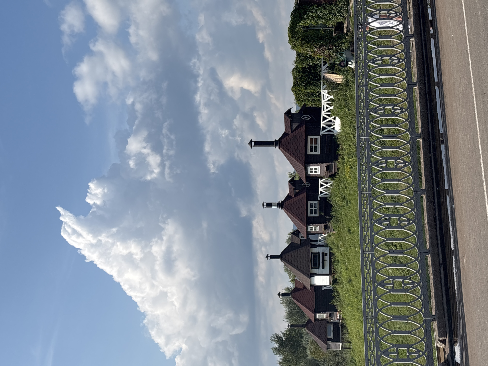 The image depicts a picturesque scene in Naberezhnye Chelny, Russia, featuring a row of distinctive dark-colored cabins with conical, dark red roofs, each topped with a chimney. These cabins are situated on a grassy incline, suggesting a waterside location or a resort area. A decorative white fence runs along the front of the cabins, adding a touch of quaintness. In the foreground, a grey ornate metal railing separates a road from the grassy embankment where the cabins are located. The sky above is a vibrant blue, punctuated by dramatic, billowing white clouds, creating a dynamic and aesthetically pleasing atmosphere. The overall impression is one of tranquility and natural beauty, with a hint of rustic charm offered by the unique architecture of the cabins.