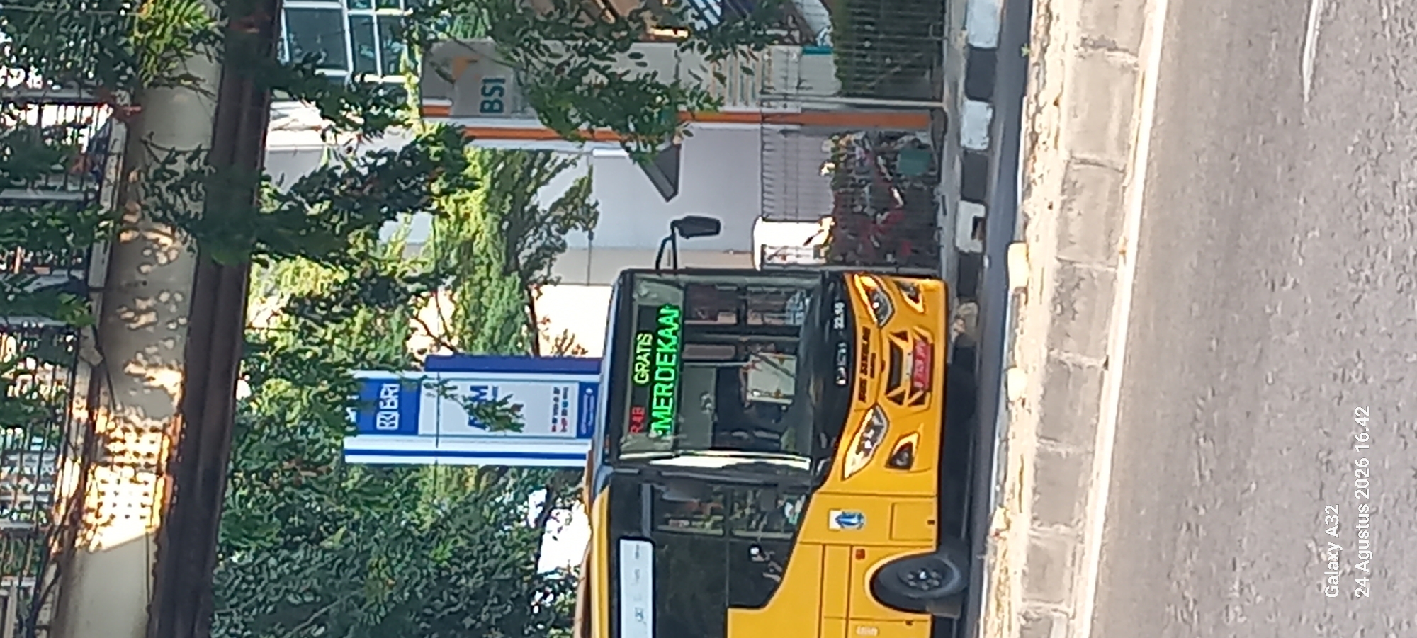 A yellow city bus is parked on the side of a street in North Jakarta, Indonesia. The bus has its destination display lit up with green text that reads "R.48 GRATIS MERDEKAAN". Behind the bus, a tall blue sign with the BRI logo and "ATM" is visible. To the right of the BRI sign, another blue and white sign with "BSI" is present. The bus has its headlights on and appears to be in traffic. The street has a concrete median and sidewalk. Trees and other foliage are visible in the background, along with part of a building. The sky is not visible. The overall scene depicts a typical urban environment with public transportation and financial institutions.