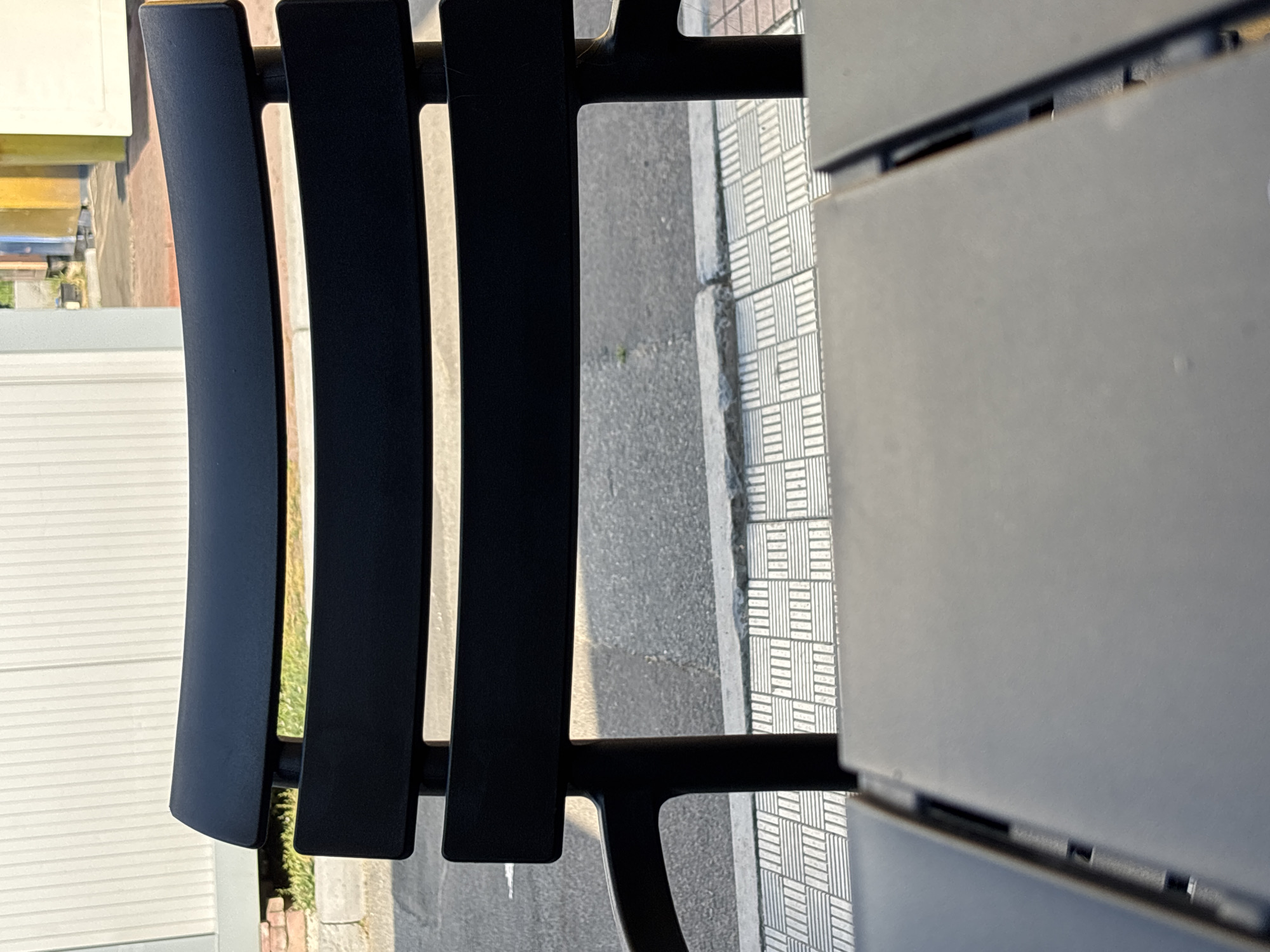 A close-up, eye-level shot shows the back of a black outdoor chair. The chair has a curved top slat and two horizontal slats below it, all made of black metal. The chair is positioned in front of a sidewalk with a repeating geometric pattern of light gray squares. Beyond the sidewalk, there is a paved road. In the background, a white corrugated wall is visible, with hints of greenery and some yellow and beige structures further in the distance. The lighting suggests it is daytime, with shadows casting on the ground. The foreground is dominated by a dark gray surface, likely a table, partially obscuring the view of the chair's legs. There are no people or animals present in the scene.