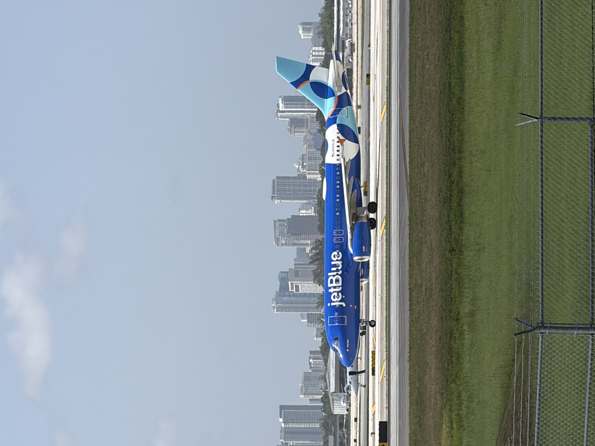 A JetBlue airplane is on the tarmac at an airport, with a city skyline in the background. The airplane is bright blue with the JetBlue logo on its side and a colorful design on its tail. It is a sunny day with a light blue sky and a few clouds. The airplane is positioned to the right of the frame, facing left, with its landing gear deployed. In the background, a dense cluster of modern high-rise buildings forms the city skyline. In the foreground, there is a grassy area and a chain-link fence. The image appears to have been taken in Dania Beach, Florida, given the distinctive skyline which is consistent with Fort Lauderdale's skyline, located in close proximity to Dania Beach.