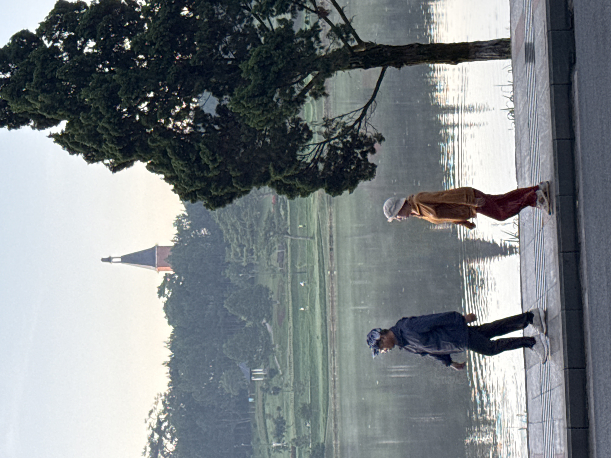 The image depicts Xuan Huong Lake in Da Lat, Vietnam, at what appears to be sunrise or sunset due to the soft lighting. Two individuals are walking on the sidewalk that borders the lake. On the left, a man wearing a dark blue jacket, dark pants, and a patterned baseball cap is walking towards the right of the frame. He has white athletic shoes on. To his right, a woman wearing a light brown jacket, red pants, and a light-colored brimmed hat is also walking towards the right. She also has athletic shoes on. Behind them, the calm surface of Xuan Huong Lake reflects the sky and the surrounding lush green landscape. In the background, a prominent clock tower, the Dinh 2, stands above a dense line of pine trees. The scene conveys a sense of peaceful morning or evening activity.