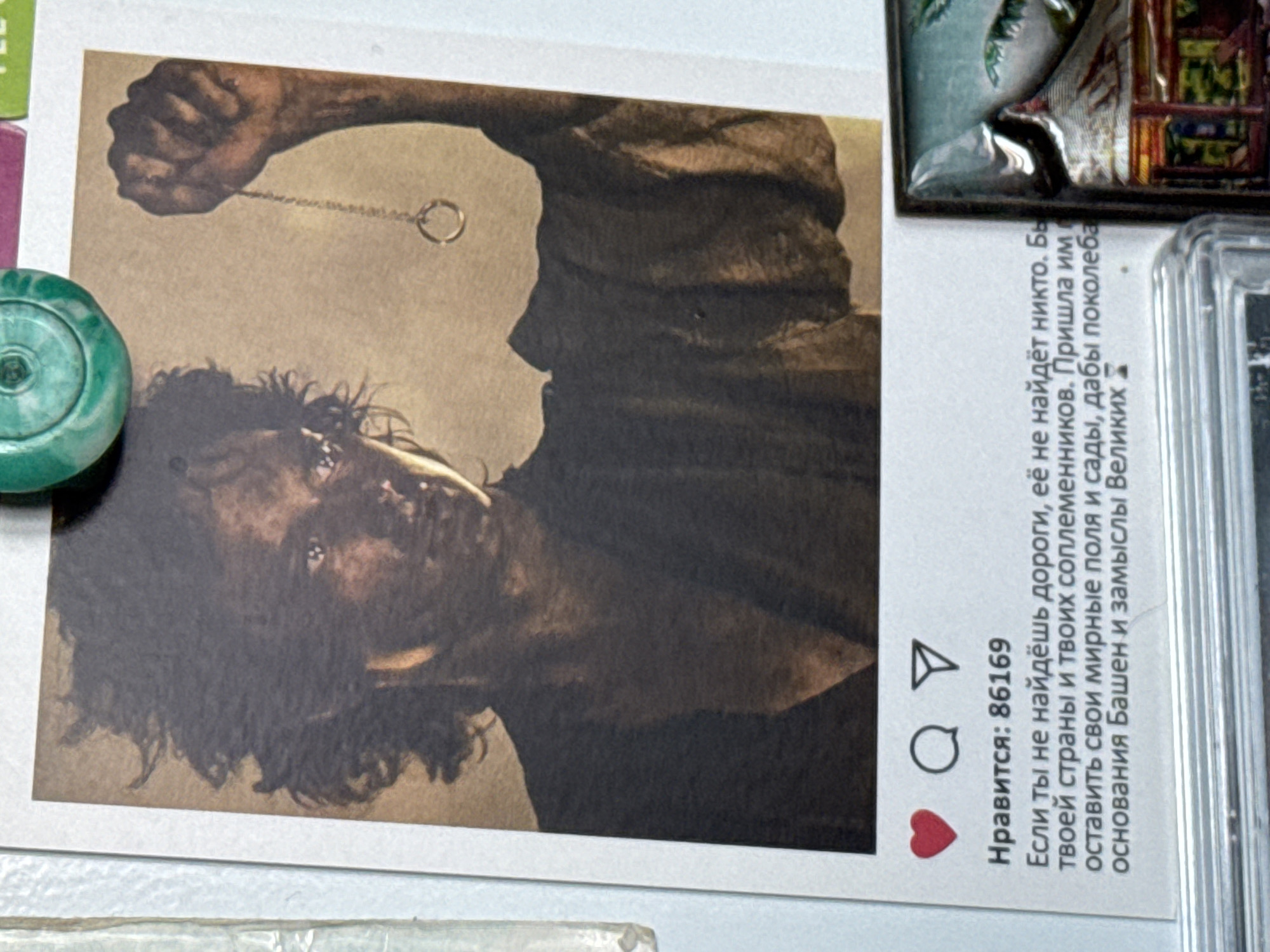 The image shows a close-up of a photograph or print of a character from a fantasy setting, likely from "The Lord of the Rings," holding a ring on a chain. The print is framed to resemble an Instagram post, complete with a heart icon, a speech bubble icon, and a mountain/paper airplane icon. Below the icons, the text "Нравится: 86169" (Likes: 86169) is visible, followed by a caption in Russian: "Если ты не найдёшь дороги, её не найдёт никто. Бы твоей страны и твоих соплеменников. Пришла им оставить свои мирные поля и сады, дабы поколеба основания Башен и замыслы Великих" (If you do not find the way, no one will find it. To your country and your kin. They came to leave their peaceful fields and gardens, to shake the foundations of the Towers and the plans of the Great).
The print is attached to a white, textured surface, possibly a refrigerator or a magnetic board. A green, circular object, possibly a magnet or decoration, is visible at the top of the image, partially obscuring the print. To the right of the print, a portion of another object, possibly a framed picture or souvenir with a tropical scene, is visible. The overall setting appears to be indoors. The image does not contain any visual information to suggest a specific location within the city of Izhevsk, Russia.