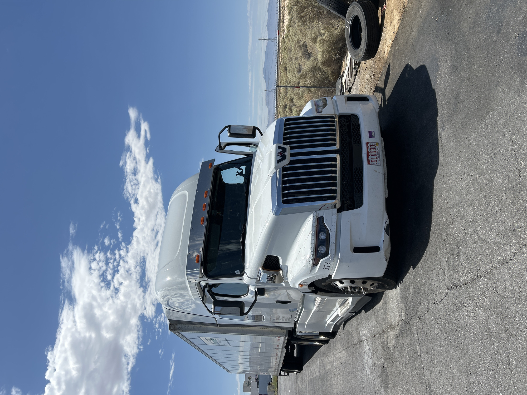 A white semi-truck is parked on a cracked asphalt lot under a bright blue sky with scattered white clouds. The truck has a chrome grille and a trailer attached. The trailer has "Premier" written on its side. A license plate on the front of the truck reads "BL0088 COLORADO". To the right of the truck, several large tires are stacked on the ground, and a chain-link fence encloses an area with dry brush and distant mountains. In the background, power lines and towers are visible. The scene appears to be in an industrial or service area.