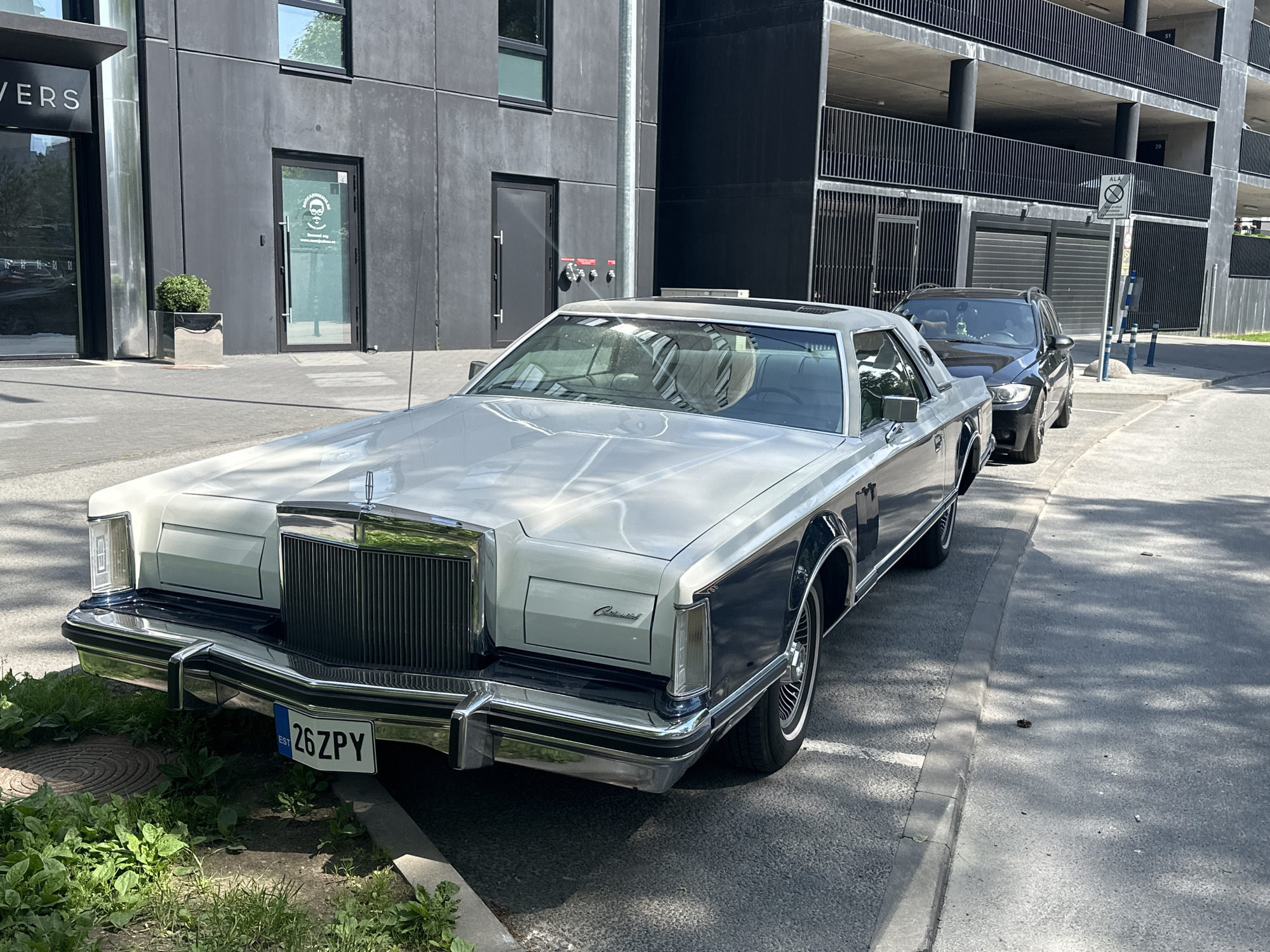 A two-tone white and dark blue 1970s Lincoln Continental Mark V is parked on a street in Tallinn, Estonia. The car is positioned on the right side of the frame, with its front end facing the viewer. A dark blue BMW SUV is parked behind it. The background shows a modern, multi-story building with a dark facade and a covered parking structure. The car has a license plate that reads "26 ZPY". The scene is illuminated by sunlight, casting shadows on the ground and the cars.