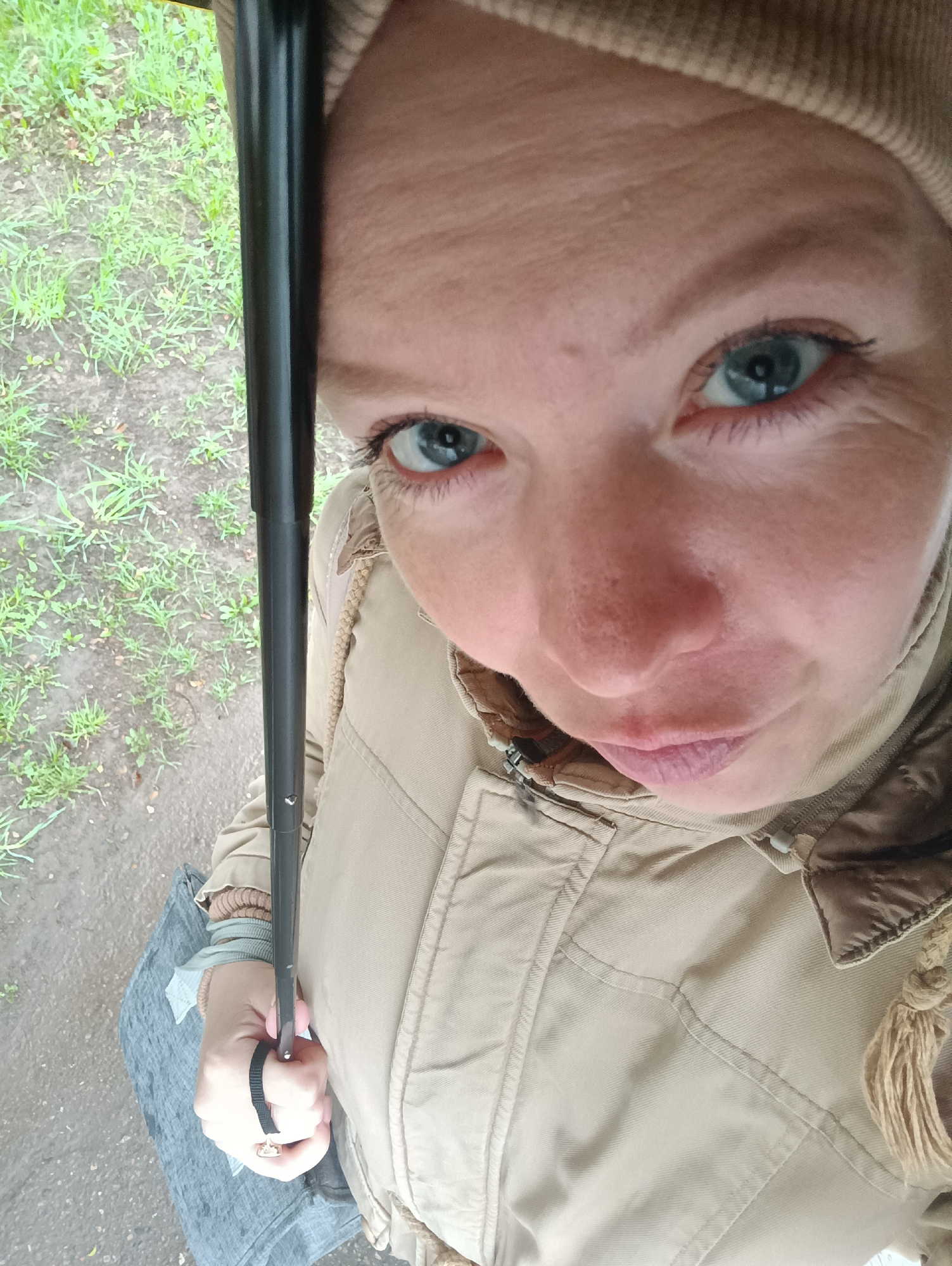 A close-up, high-angle shot depicts a fair-skinned person with blue eyes, wearing a beige hat and jacket, holding a black umbrella. The lower part of the scene shows a gray, possibly wet, surface, possibly a sidewalk or road, with scattered green vegetation. The location is unspecified, but the city mentioned is Chaikovsky, Russia.