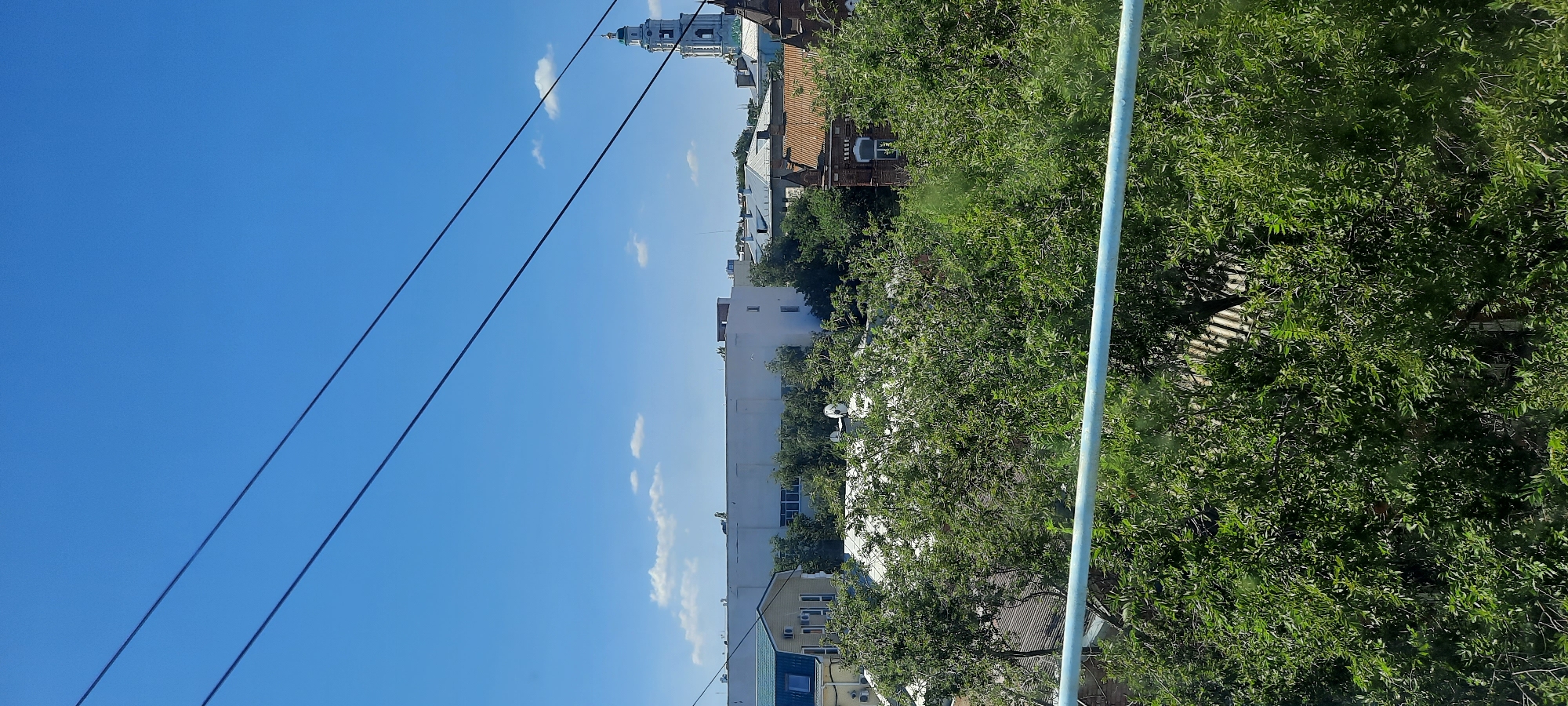 The image captures a view of a cityscape under a bright blue sky with scattered white clouds. Three dark, thin wires run diagonally across the upper portion of the frame. In the middle ground, a variety of buildings are visible, including a long, white, flat-roofed building and a smaller, light yellow building with a dark green roof. Trees with lush green foliage partially obscure some of the structures. On the right side of the image, a tall, white bell tower with a green dome and spire rises above the other buildings, its architectural style suggests it is likely part of a church or cathedral. This bell tower is characteristic of the architectural landscape of Astrakhan, Russia, where numerous religious structures with similar features are found. In the foreground, a light blue railing or pipe is visible, suggesting the photograph was taken from an elevated position. The overall scene depicts a typical urban environment in Astrakhan on a sunny day, with a blend of modern and historic architecture, surrounded by abundant greenery. There are no people or animals visible in the frame.