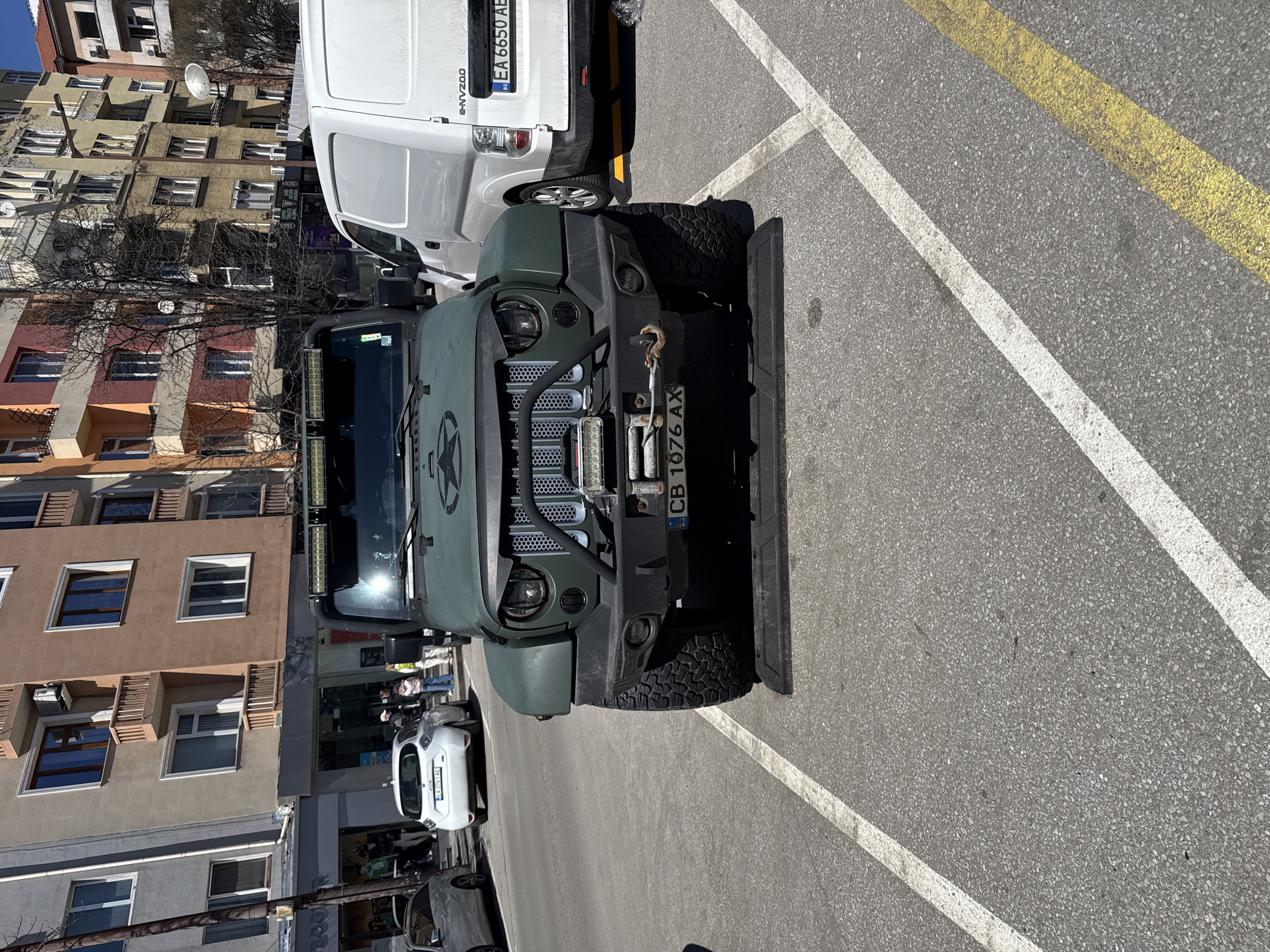 The image shows a green modified Jeep Wrangler parked in a designated parking spot in Sofia, Bulgaria. The Jeep has an aftermarket grille, a winch, auxiliary lights, and a star emblem on the hood. The license plate reads "CB 1076 AX". A white e-NV200 van with license plate "EA 6650 AE" is parked next to the Jeep. The parking spot is marked with white lines and bordered by a yellow line. In the background, there are multi-story buildings typical of Sofia's architecture. A white Renault car is visible behind the Jeep. Pedestrians are walking by in the distance. The weather appears to be sunny.