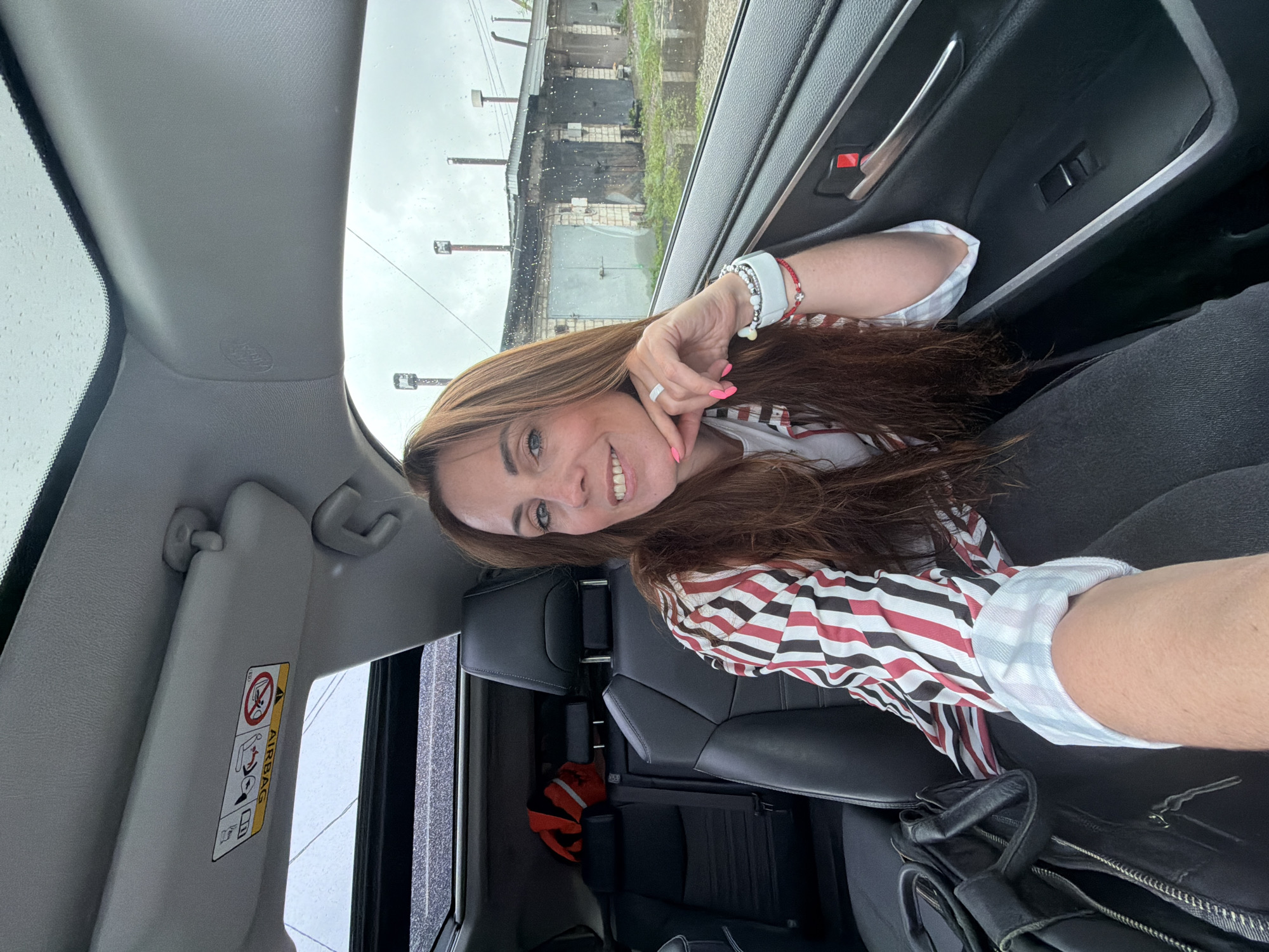 A woman with long, auburn hair is sitting in the driver's seat of a car. She is wearing a striped shirt with red, white, and black vertical stripes, and a white undershirt. Her left hand is resting on her chin, and she is smiling at the camera. She is wearing jewelry on her left wrist and a ring on her left hand. A black bag is on the passenger seat. The car has a sunroof, and the sky outside is overcast. In the background, there are garages and some industrial-looking buildings. The image appears to have been taken in Birobidzhan, Russia, given the context provided.
