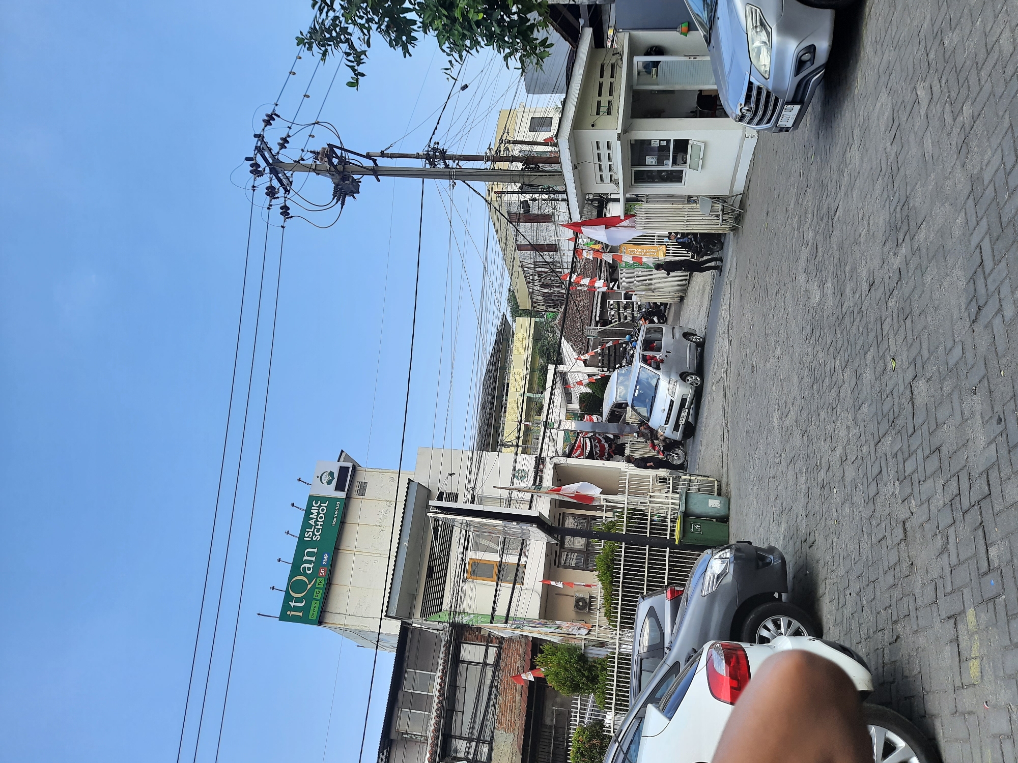 The image captures a street scene in Cimenyan, Indonesia, likely in front of the ItQan Islamic School, as indicated by a prominent green sign. The street is paved with interlocking brick pavers. Several cars are parked or moving on the street, including a silver Toyota Innova in the foreground right and a white car on the left foreground. A small silver van is seen moving down the street. A guard booth or small building is present on the right side of the road. Indonesian flags are displayed on poles and attached to buildings, suggesting a national celebration or commemoration. Various trees and foliage are visible, as are power lines and a utility pole. The sky is clear and blue. The image appears to have been taken during daylight hours.