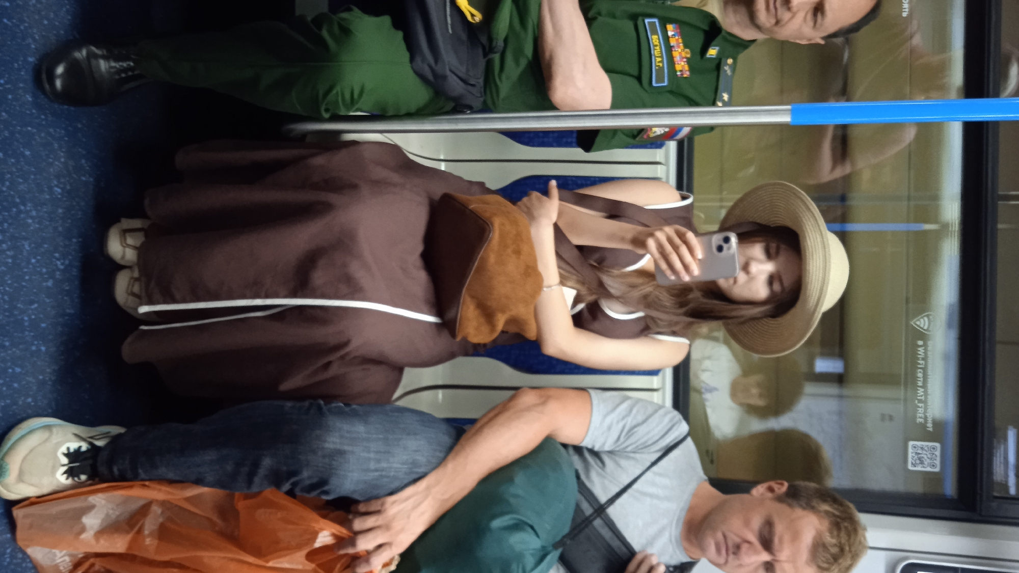 In an indoor setting with blue flooring, two individuals are seated next to each other. The person on the left is wearing dark trousers, a black shoe, and a green uniform with medals and insignia. Their legs are bent, with the left leg extending forward. Beside them, a person in a brown dress and a straw hat is holding a phone in their right hand, seemingly taking a selfie. They are also wearing a bracelet on their left wrist. The individual on the right is wearing a grey t-shirt and dark trousers. Their right arm is visible, resting on a green cushion. An orange plastic bag lies at their feet. A black bag with straps is visible on their shoulder. Reflections in a glass partition show the faces of the people.