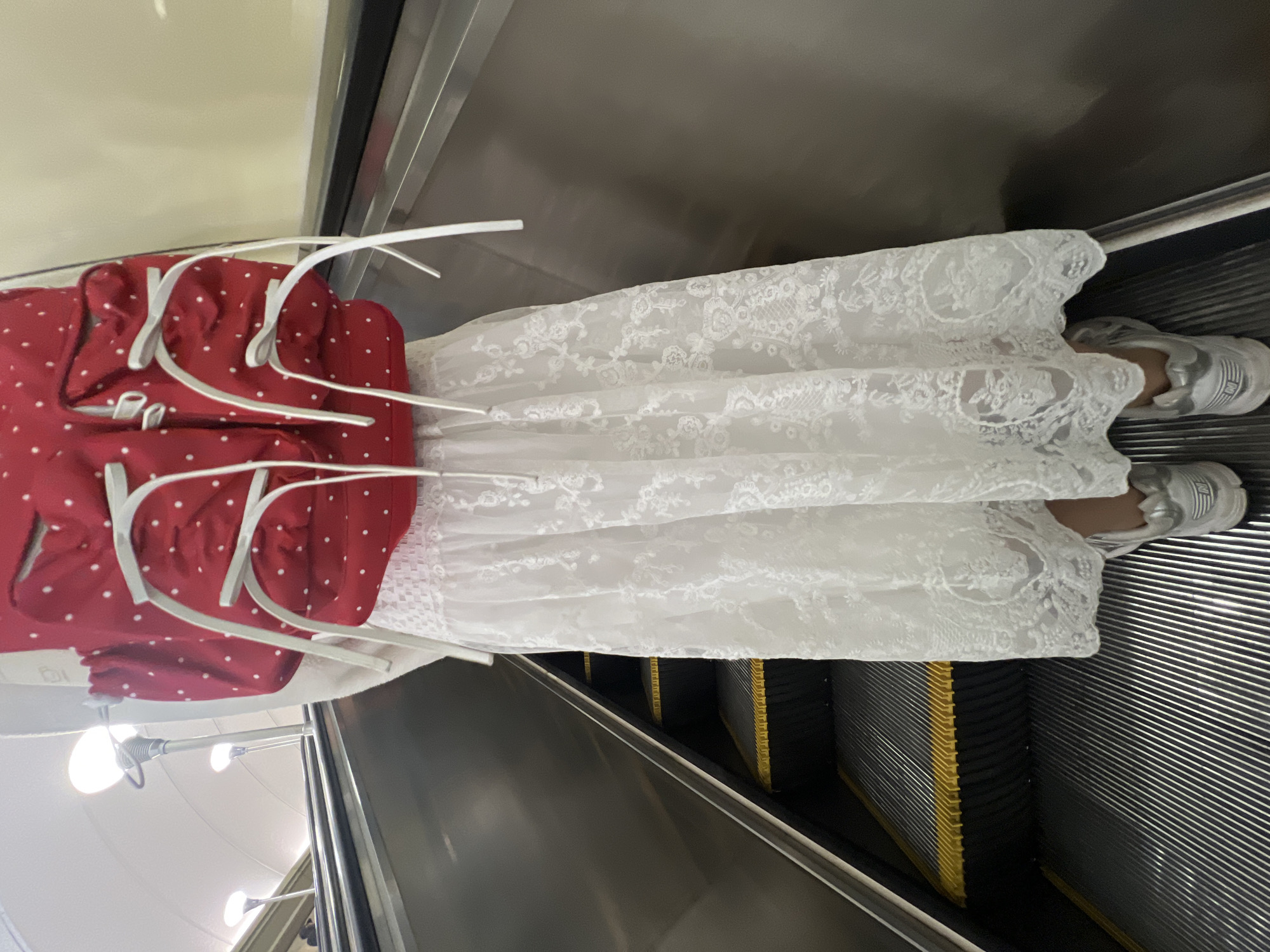 A person wearing a white lace skirt and gray sneakers is standing on an escalator, with a red and white polka-dot jacket hanging on a hanger beside them. The escalator is metallic with yellow accents. The location appears to be indoors, potentially in Saint Petersburg, Russia, based on the architecture visible in the background.