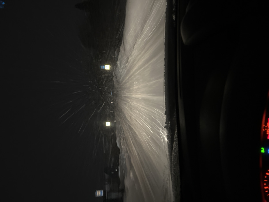 The image displays a night-time perspective from inside a vehicle on a snow-covered road, likely in Davydkovo, Russia. The foreground exhibits the dark dashboard and the lower edge of the windshield, with faint red and green illuminated indicators visible at the very bottom left. Heavy snowfall is actively illuminated by the vehicle's headlights, creating numerous horizontal streaks of light emanating forward, appearing as a dense, radial pattern against the dark background. The road ahead is entirely covered in a thick layer of snow, with accumulated snow forming banks along the sides. In the mid-distance, two distinct square-shaped, yellow-white illuminated objects or signs are visible, positioned symmetrically towards the left and right of the central axis of the road. Further to the left background, a vertical blue and white structure or sign is partially discernible. The sky is dark and overcast by night and snowfall. No individuals are visible, but the perspective indicates an occupant is present within the vehicle.