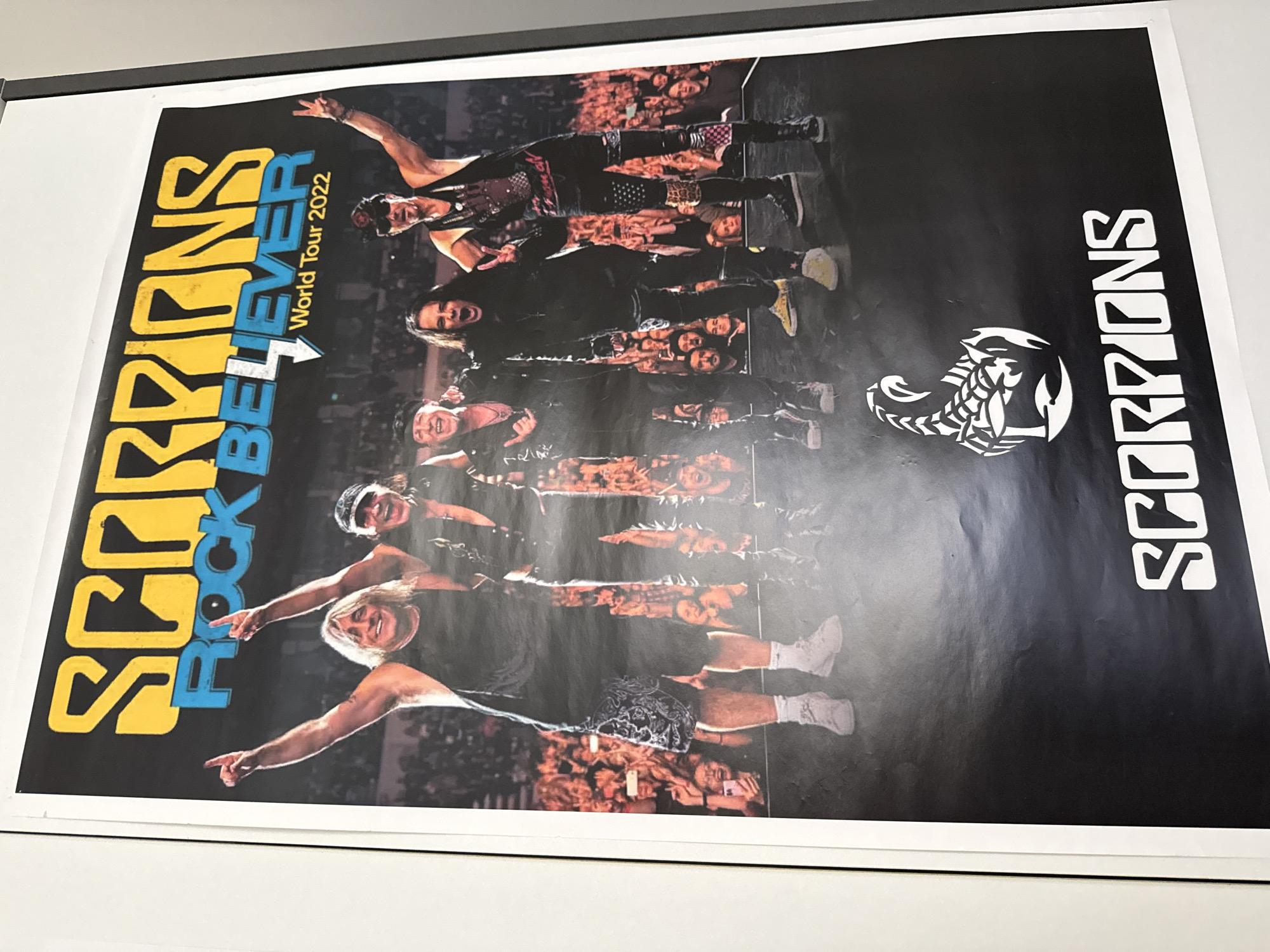 This is a close-up, angled photograph of a poster for the band Scorpions' "Rock Believer World Tour 2022." The poster features a live band photo with the band members on stage in front of a crowd. The band's name, "SCORPIONS," is prominently displayed in large yellow and blue lettering at the top, followed by "ROCK BELIEVER" and "World Tour 2022." Below the band members, a white scorpion logo is depicted. The poster is framed by a white border and the dark frame of a display. The poster is mounted on a wall, and the photograph is taken from a slightly elevated position, looking down. The text on the poster is legible. The image does not contain any information about the location in the city of Bad Sachsa, Germany.