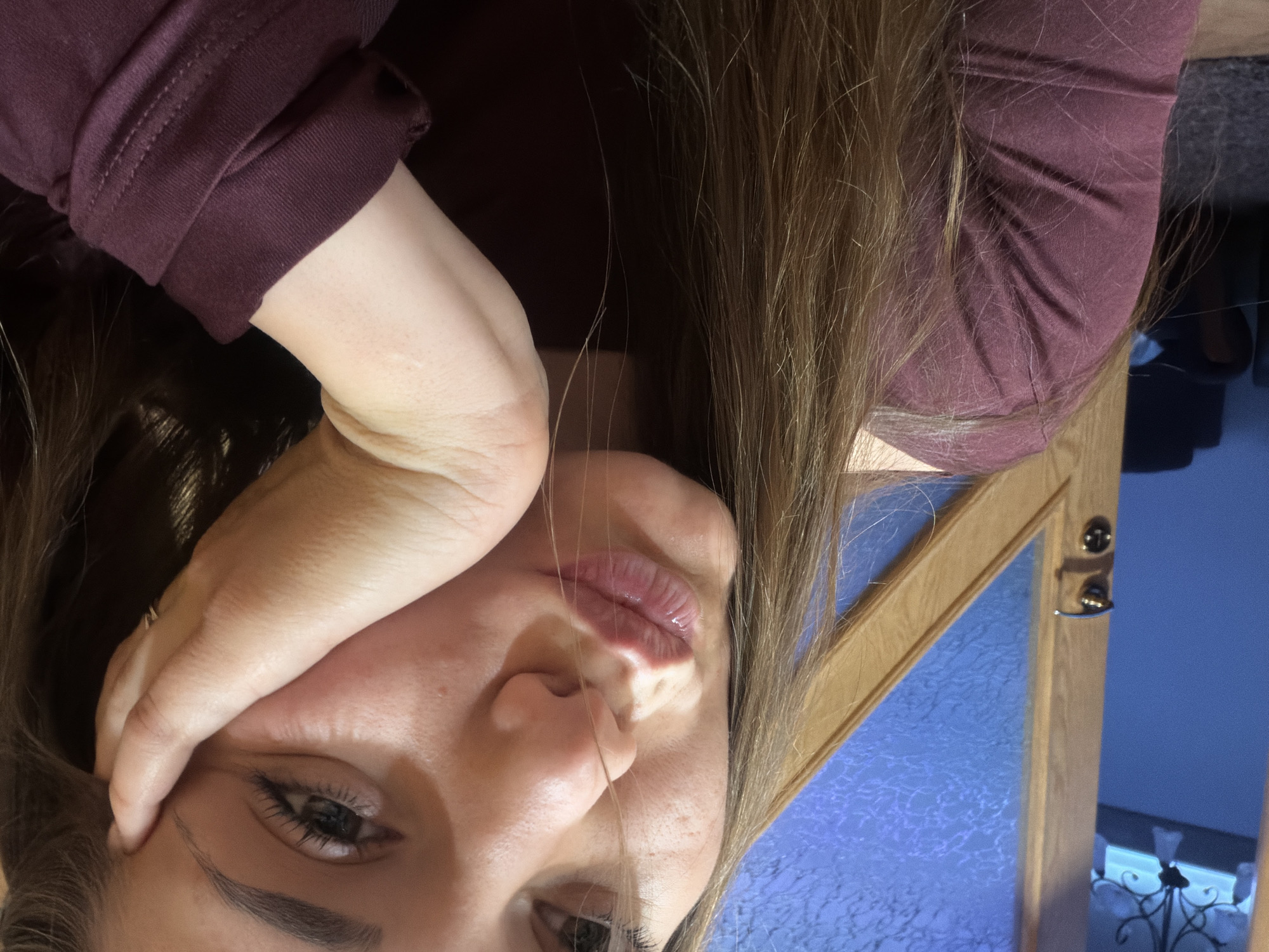 A close-up, rotated portrait of a woman. She has light skin, brown hair, and wears a burgundy top. Her right hand is touching her forehead, obscuring part of her face. A wooden framed mirror with a textured blue surface is visible to her right. The environment suggests an indoor setting. There is no information to determine the location is in Buhovo, Bulgaria.