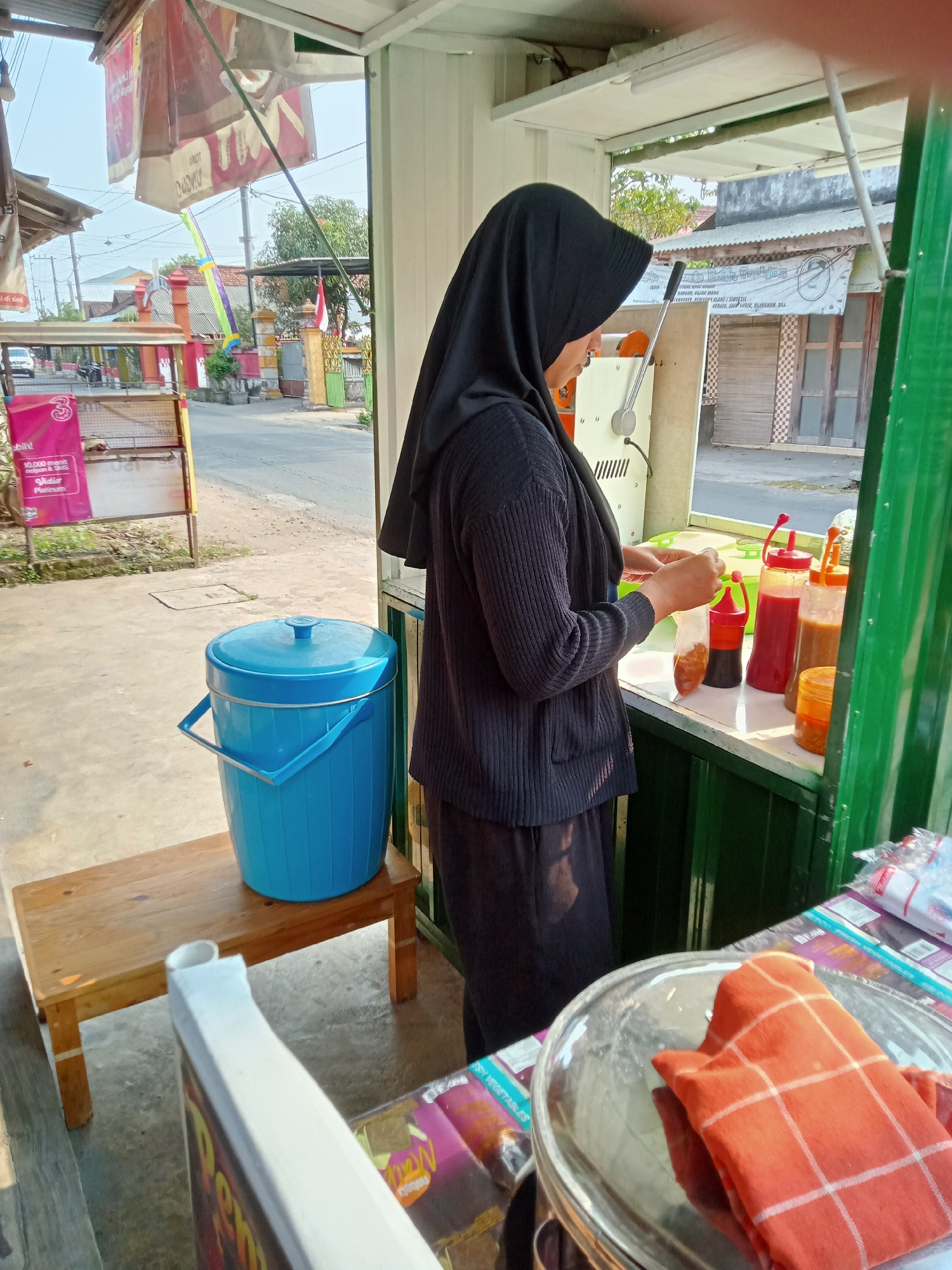 A woman wearing a black hijab and black clothing stands at a food stall in Simo, Indonesia. She is shown from the waist up, facing the right of the frame, and is in the process of preparing a food order. She holds a clear plastic bag in her left hand, which contains a brown, granular substance. Her right hand is shown reaching into the bag, seemingly to adjust its contents or close it. In front of her, on a counter, are several condiment bottles: a red bottle, a dark brown bottle, and a lighter brown bottle, along with a small orange jar. The stall appears to be open-air, with a green metal frame visible on the right. To the left of the stall, a bright blue plastic cooler with a lid and handles sits on a simple wooden bench. In the background, a street is visible, with buildings and trees lining it. The overall lighting suggests it is daytime, with sunlight casting shadows.