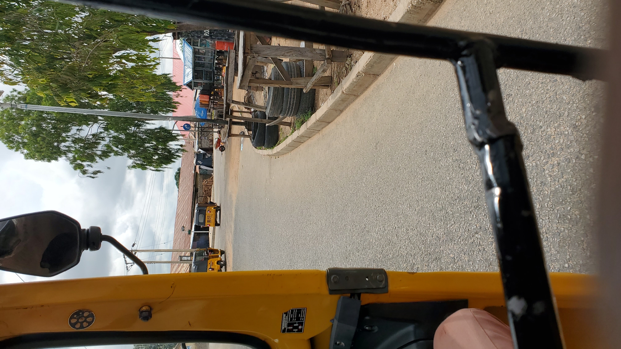 The image depicts a street scene in Jalingo, Nigeria, likely taken from the interior of a yellow auto-rickshaw. In the foreground, the yellow body of the auto-rickshaw is visible on the left, along with a black metal bar. The road is paved and appears to be asphalt.
Further down the road, two auto-rickshaws are driving away from the camera. The one on the left is yellow, and the one on the right is yellow with a black roof and features the license plate "359T O".
On the right side of the road, there are several wooden stalls with rough wooden tables, some of which are supported by stacked tires. Behind the stalls, there are buildings with corrugated metal roofs, and a large tree with green foliage dominates the background. The sky is cloudy. The overall impression is that of a typical urban or semi-urban street environment in Nigeria.