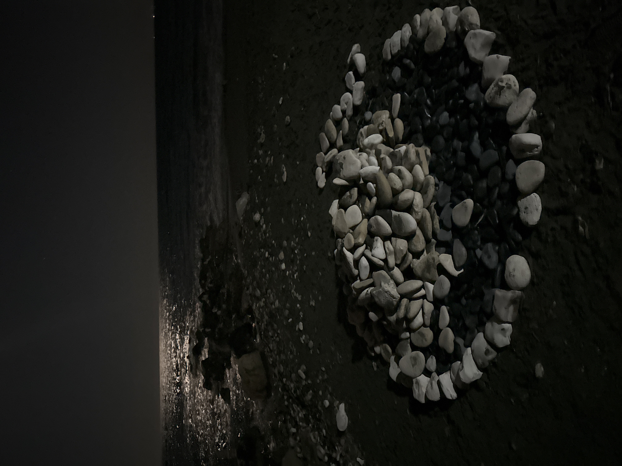A nighttime scene at a beach in Agios Tychona Community, Cyprus. The image captures a dark, moonlit ocean with gentle waves lapping at a shore covered in dark sand and pebbles. In the foreground, a large arrangement of light-colored, smooth stones is piled in the center, forming a rough spiral shape extending outwards. The stones are of various sizes and shapes, with some reflecting the moonlight. The surrounding beach is also scattered with smaller pebbles. The sky is dark and clear, with a faint glow on the horizon from the moon reflecting on the water.