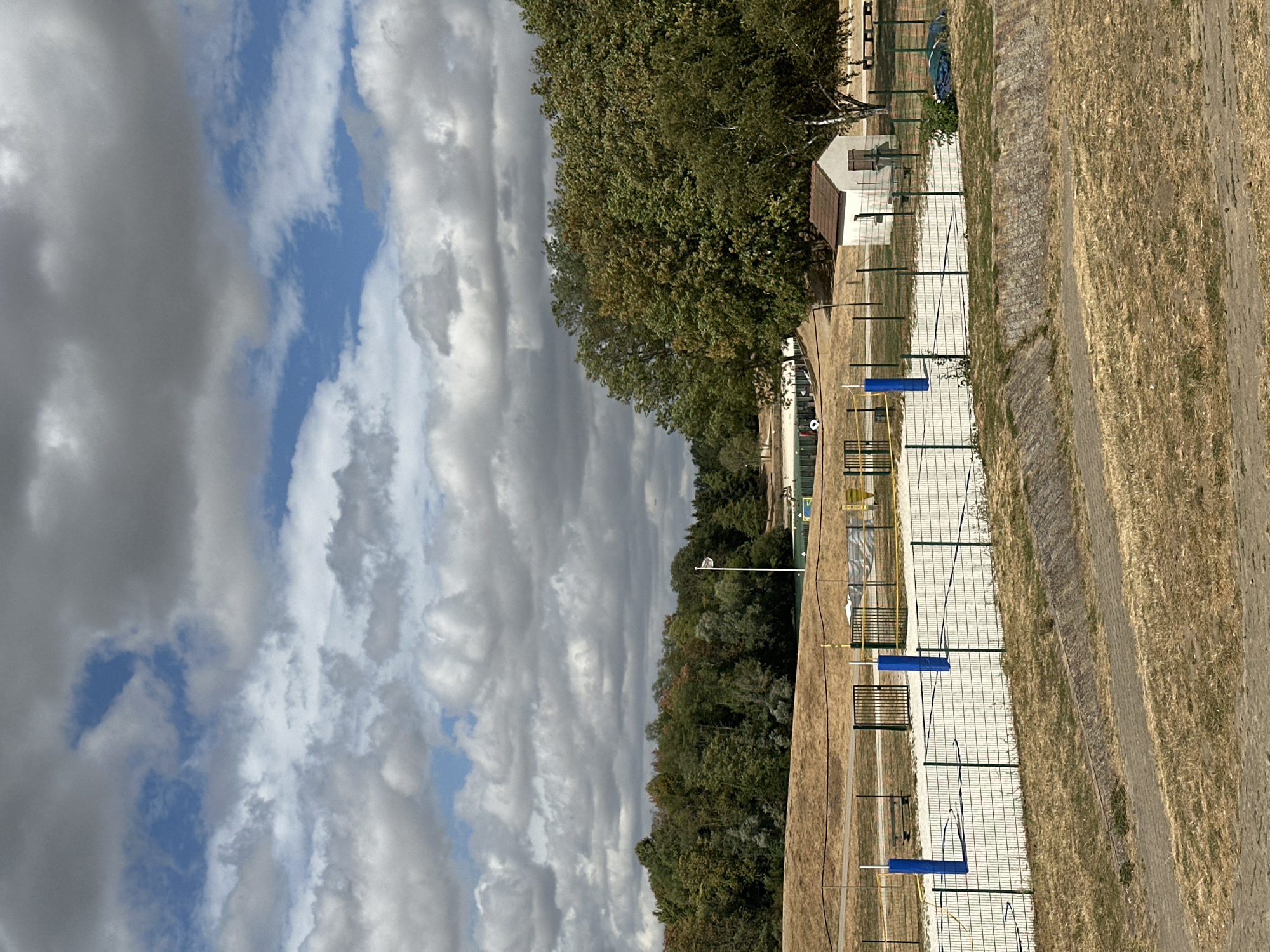 This is a high-angle, full-shot photograph of an outdoor recreational area in Jablines, France, on a cloudy day. In the foreground, dry, golden grass covers a sloping embankment. A stone retaining wall separates this area from a paved pathway. Beyond the pathway is a fenced-in sand court, likely for volleyball, with two tall blue posts at the net line. A green metal fence surrounds the sand court. In the middle ground, a small white building with a brown roof sits to the right of the sand court. Further back, a line of green trees forms a dense forest. A white flag on a flagpole stands in the distance. In the background, more trees are visible, and beyond them, a building with a light-colored roof and white walls can be seen. The sky is filled with a mix of dark grey and white clouds, with patches of blue sky showing through.