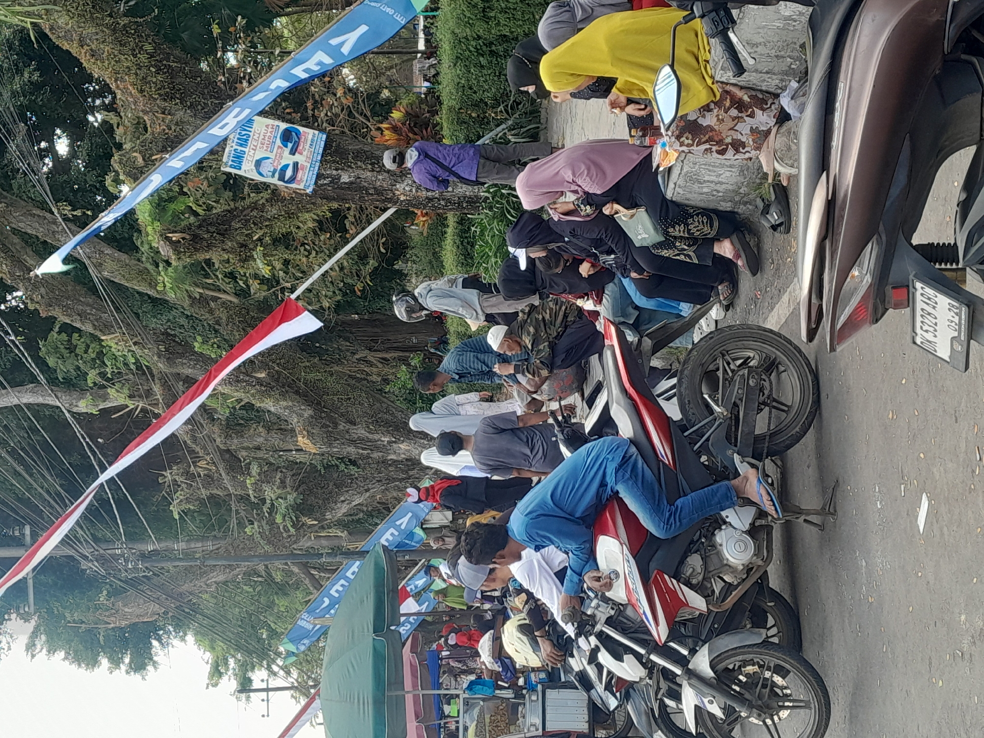 The image depicts an outdoor scene likely in Genteng Kulon, Indonesia, with a group of people gathered, several motorcycles, and a street lined with trees. In the foreground, a person in a blue shirt and jeans is sitting on a red and white motorcycle. To the left of this motorcycle, another motorcycle is partially visible. A row of people, many wearing headscarves and traditional Islamic clothing, are seated on a low concrete barrier. One person wears a bright yellow headscarf, and another a pink headscarf. Behind the seated group, more people are standing and walking, including individuals in religious attire.
Several red and white Indonesian flags are displayed prominently, attached to poles and strung across wires. Blue flags with text are also visible, one of which shows an advertisement for "BANG HASYIM CENTENG SEMUA MURAH" with images of what appear to be mattresses or beds. A green awning shades a small food stall on the left side of the image, where a person is interacting with a vendor. The background features a dense line of mature trees with thick trunks and branches, suggesting a park or tree-lined street. Power lines crisscross the upper portion of the image, running between poles. The ground is paved with asphalt. The overall atmosphere suggests a public gathering or market setting on a clear day.