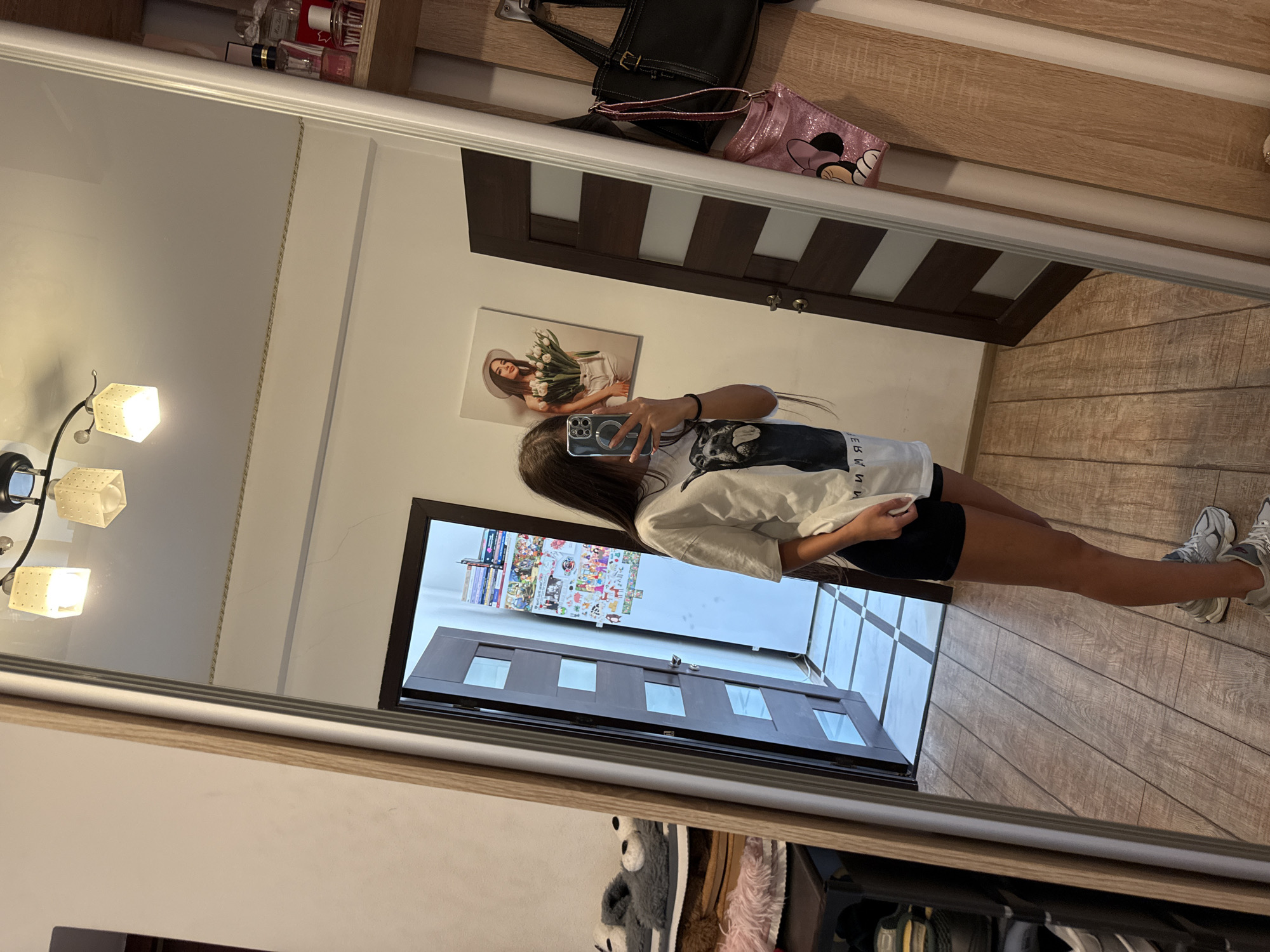 The image shows a young woman taking a mirror selfie in what appears to be a bedroom. She is wearing a white t-shirt with a graphic print and black shorts. She is holding a smartphone in her right hand and is wearing sneakers. In the background, there is a doorway with a modern design, a picture on the wall of a woman holding tulips, and shelves with various items, including perfume bottles and bags. The flooring is wooden. The overall impression is of a casual, domestic setting. There is no information in the image that indicates it is located in Chișinău, Moldova.