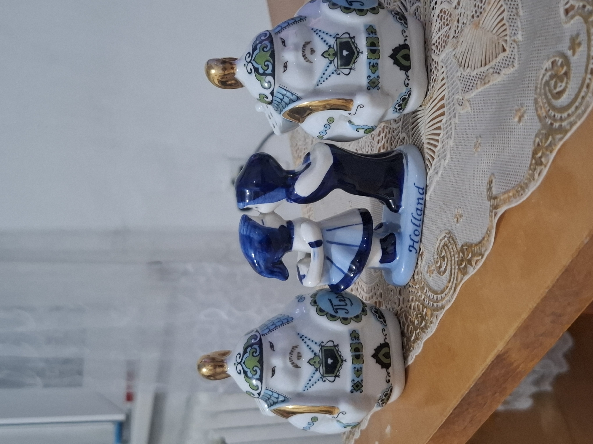 The image displays three porcelain figurines on a wooden table covered with a white lace doily. On the left and right are two identical figurines resembling small teapots, each with a golden knob on top and decorated with intricate blue, green, and black patterns. The central figurine depicts two figures, a boy and a girl, in traditional Dutch attire, kissing. The boy wears a blue and white outfit, while the girl is in a dark blue dress. The base of the central figurine has the word "Holland" inscribed on it in blue lettering. The background appears to be a blurred interior with white curtains. The lighting suggests it is daytime, likely indoors. There is no identifiable indication of the location being in Almaty, Kazakhstan.