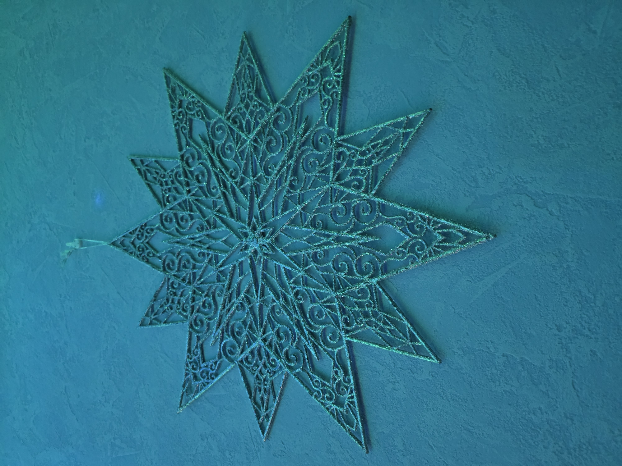 This media file showcases a turquoise, glittery, eight-pointed star ornament hanging against a textured turquoise wall. The ornament has an intricate, lacy design with swirling patterns and geometric shapes. A small, white ribbon is attached to one of the star's points, suggesting it is meant to be hung. The lighting creates a slightly dramatic effect, highlighting the glitter and the texture of the wall. The scene is static, with no activity or event taking place other than the presentation of the ornament. The overall mood is calm and decorative, possibly evoking a sense of winter or festivity. No text is visible in the image. The location context is Omsk, Russia, but there are no specific visual cues within the image to confirm this.