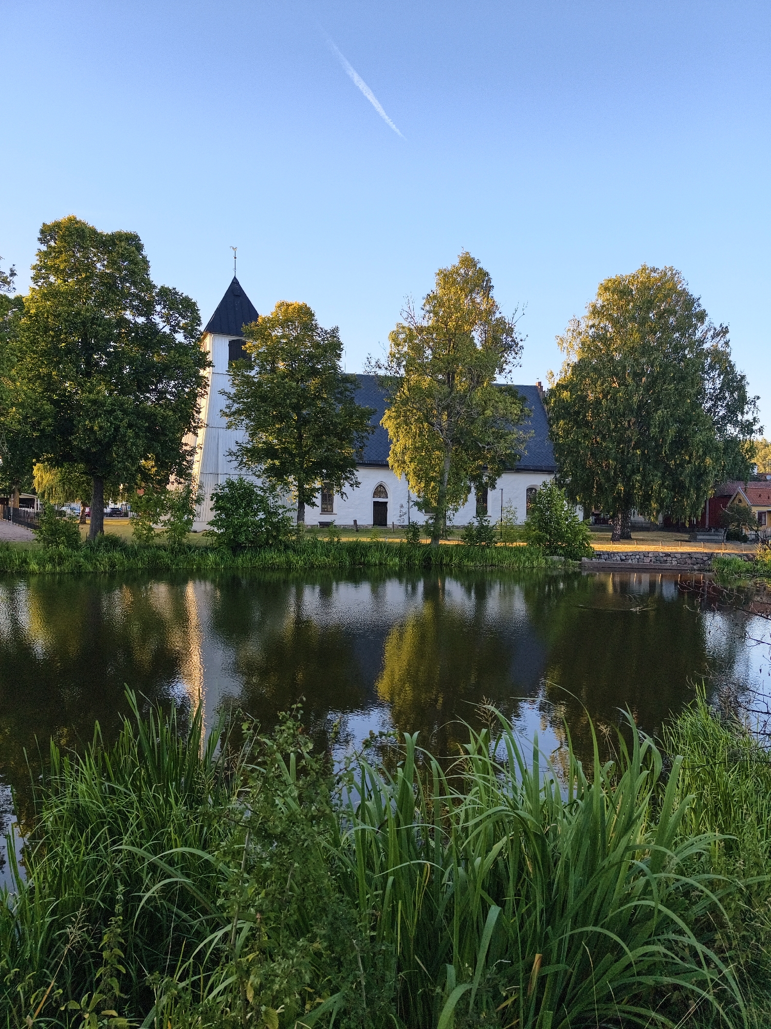 A white church with a dark pitched roof and a tall, slender tower stands on the far bank of a dark, reflective body of water. The church is surrounded by lush green trees, with some taller trees framing the building and others clustered on the right side of the image. The sky is a clear, light blue with a faint contrail visible in the upper right quadrant. In the foreground, tall, green reeds and other vegetation grow densely along the water's edge. The water itself reflects the trees and the church, creating a mirror image. The scene appears to be a serene and picturesque view of a church in a natural setting, likely within Söderköping, Sweden, given the architectural style and surrounding landscape. There are no people visible in the image, and no discernible action is taking place. The overall atmosphere is peaceful and tranquil, bathed in natural light.